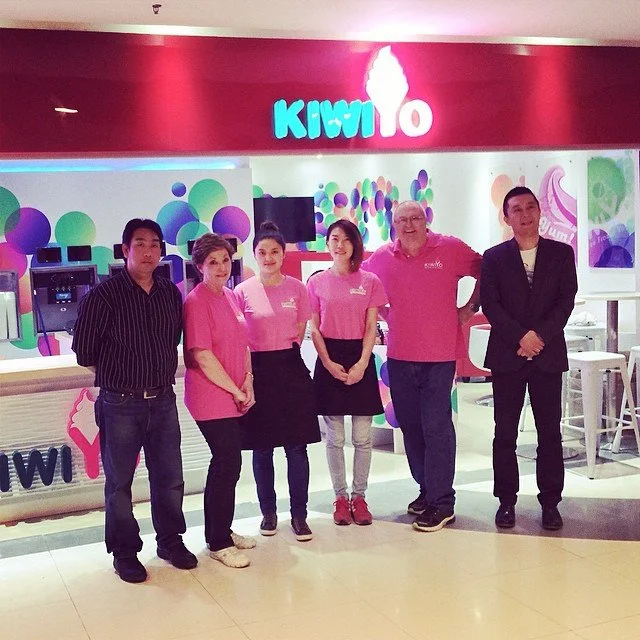 Group of six people standing in front of a Kiwi Mo frozen yogurt shop, with three employees in pink shirts and customers in casual attire.