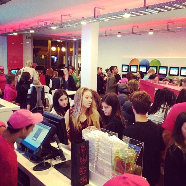 Crowded indoor space at an arcade or entertainment venue with many people, some in pink hats, a cashier station, and a row of colored computing stations along the wall.