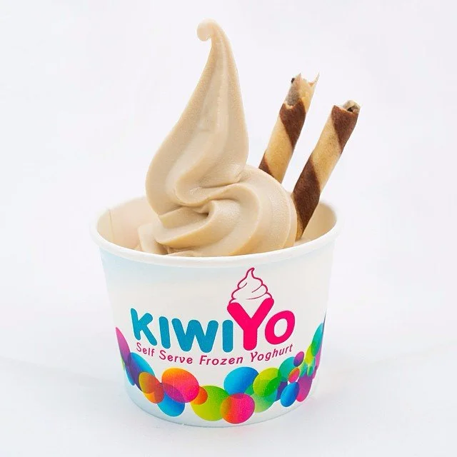 A cup of soft-serve frozen yogurt with a swirl and two chocolate-dipped wafer roll cookies, branded with KiwiYo logo.