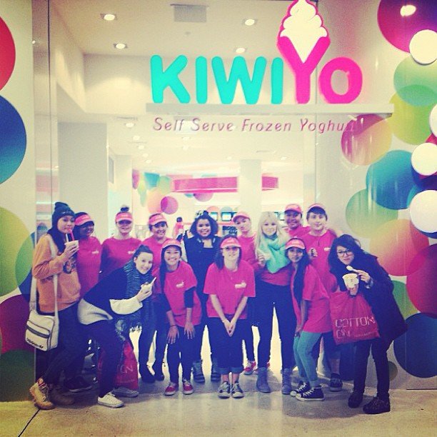 Group of people standing in front of a KIWI YO frozen yogurt shop entrance, wearing pink for a group event or outing.