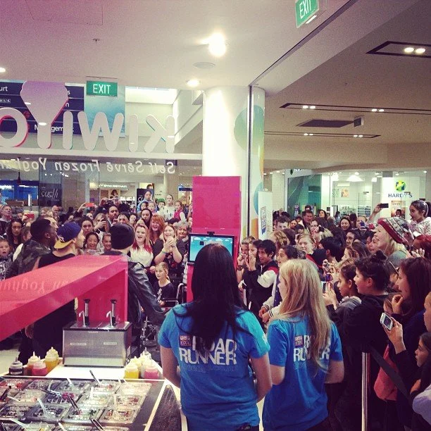 Large crowd of people inside an indoor shopping mall gathered around a food stall with two women in blue shirts. The crowd is taking photos and watching an event or presentation.