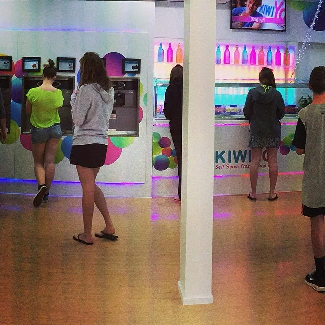 People standing in line at self-serve frozen yogurt shop with colorful bottles on display and a TV screen above