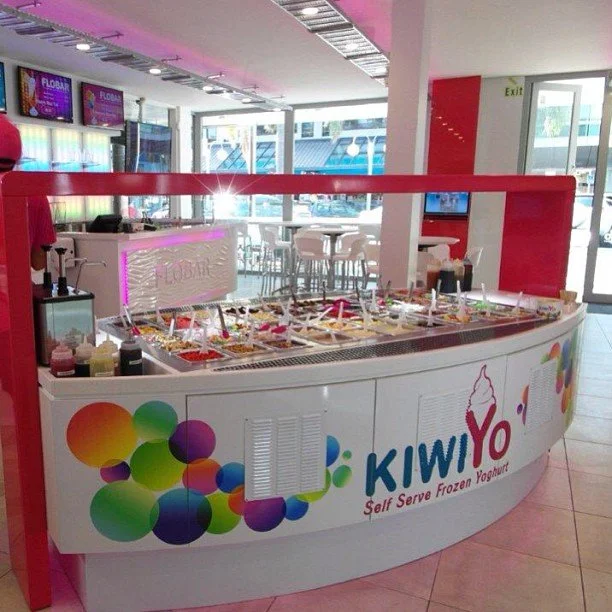 Self-serve frozen yogurt station with topping area inside a bright, modern shop, featuring colorful bubbles and a large ice cream cone graphic on the front.