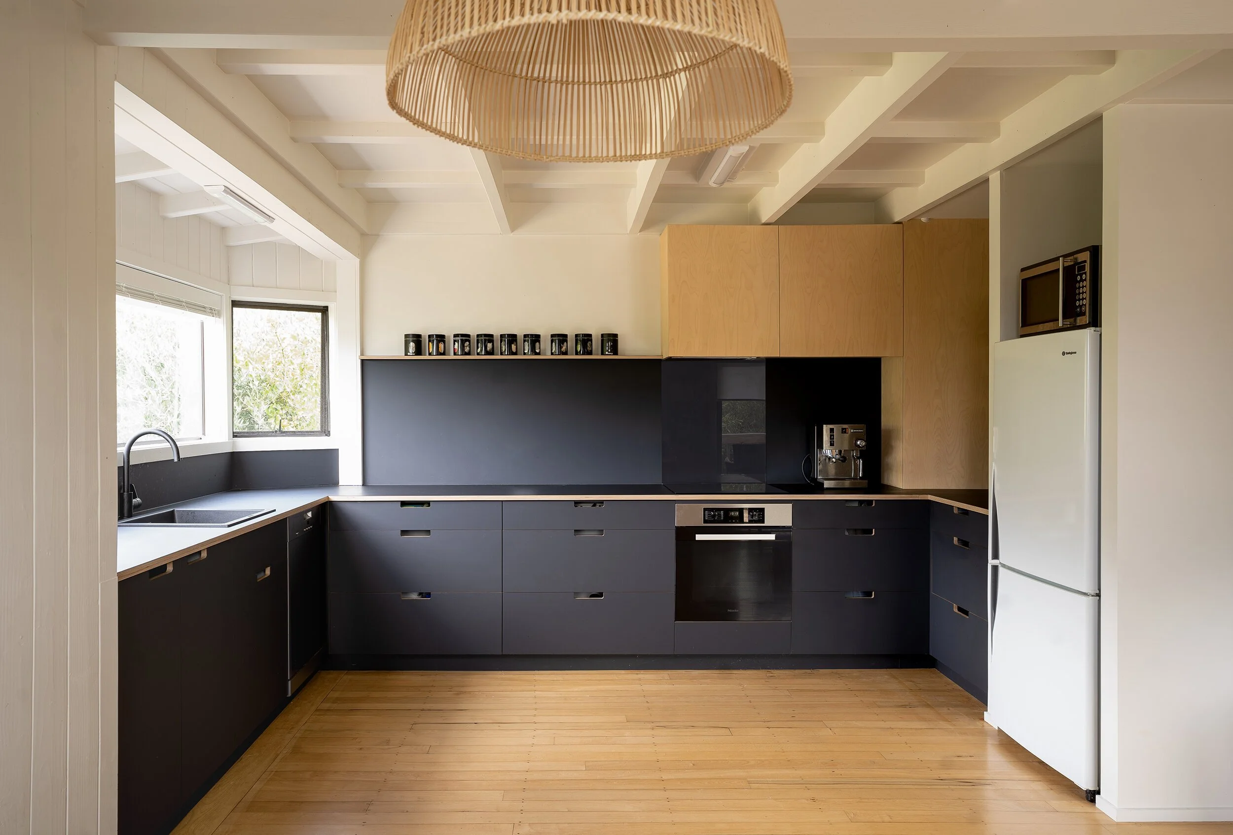 Modern kitchen with black cabinets, wooden upper cabinets, a coffee machine, and a white refrigerator. Windows above the sink, and a woven ceiling light.