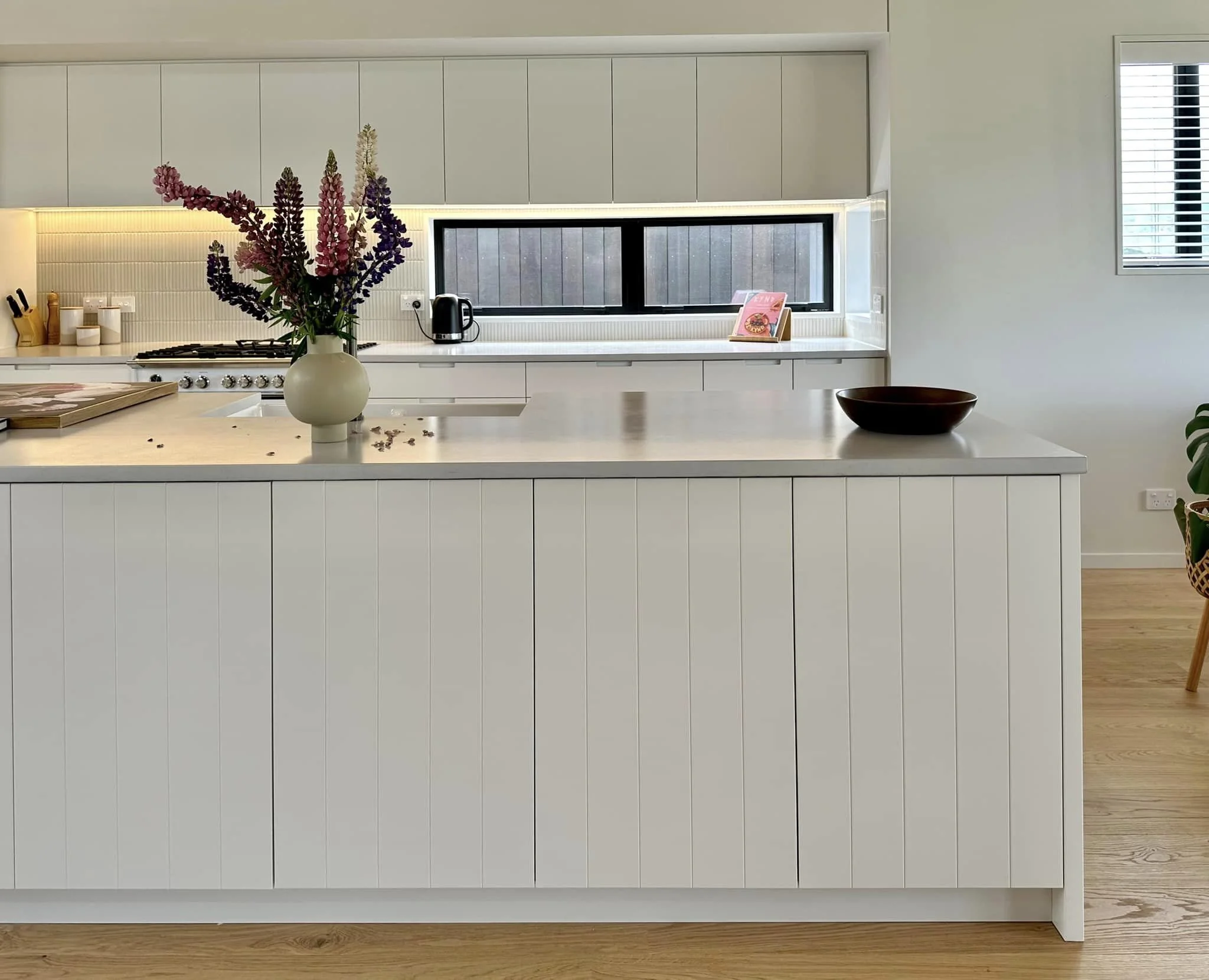 Modern kitchen with white cabinets, a white countertop, a vase with purple and pink flowers, black bowl, and a window with blinds.