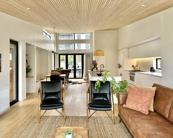 Modern open-concept living and dining area with a kitchen, featuring wooden ceiling, light-colored walls, black chairs, a brown sofa with a pink pillow, and green plants.