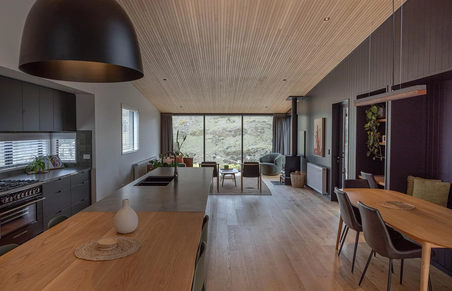 Open-concept living area with modern kitchen, dining, and sitting space featuring wood flooring, large window, and minimalist decor.