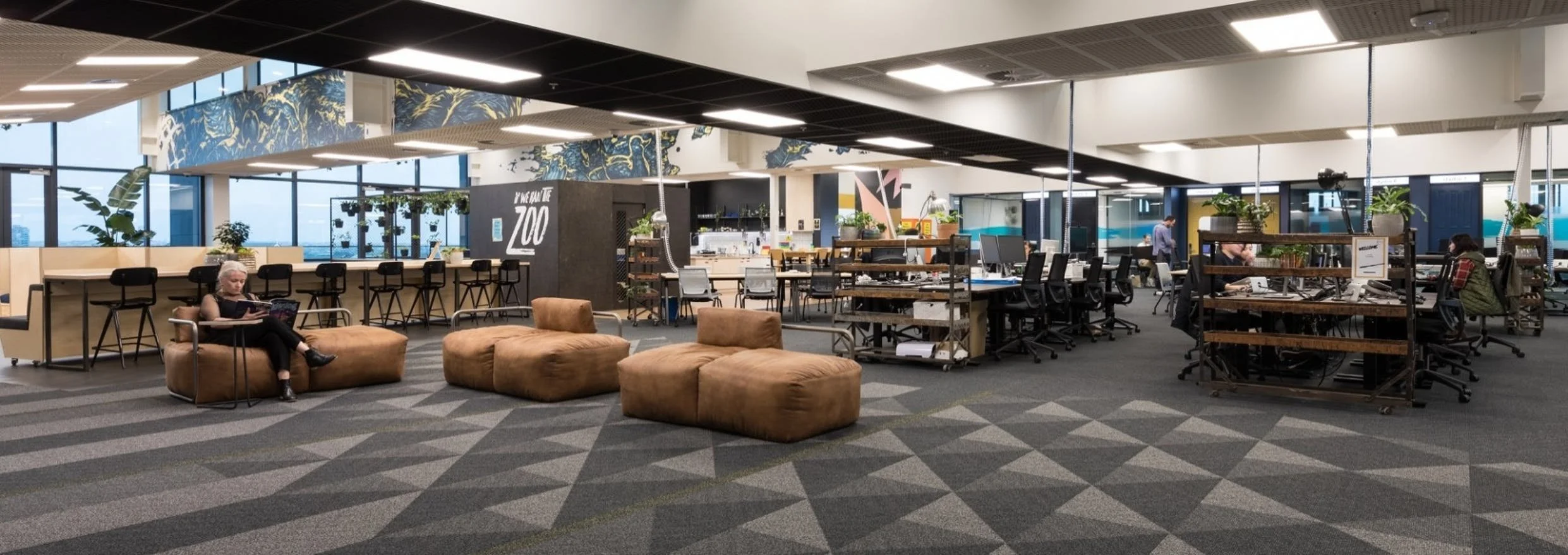 Open office space with seating areas, desks with computers, and people working, featuring large windows and modern decor.