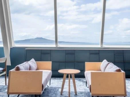 A modern interior seating area with two beige armchairs, two yellow cushions, a small round wooden table, and a large panoramic window showing an ocean view and distant mountains.