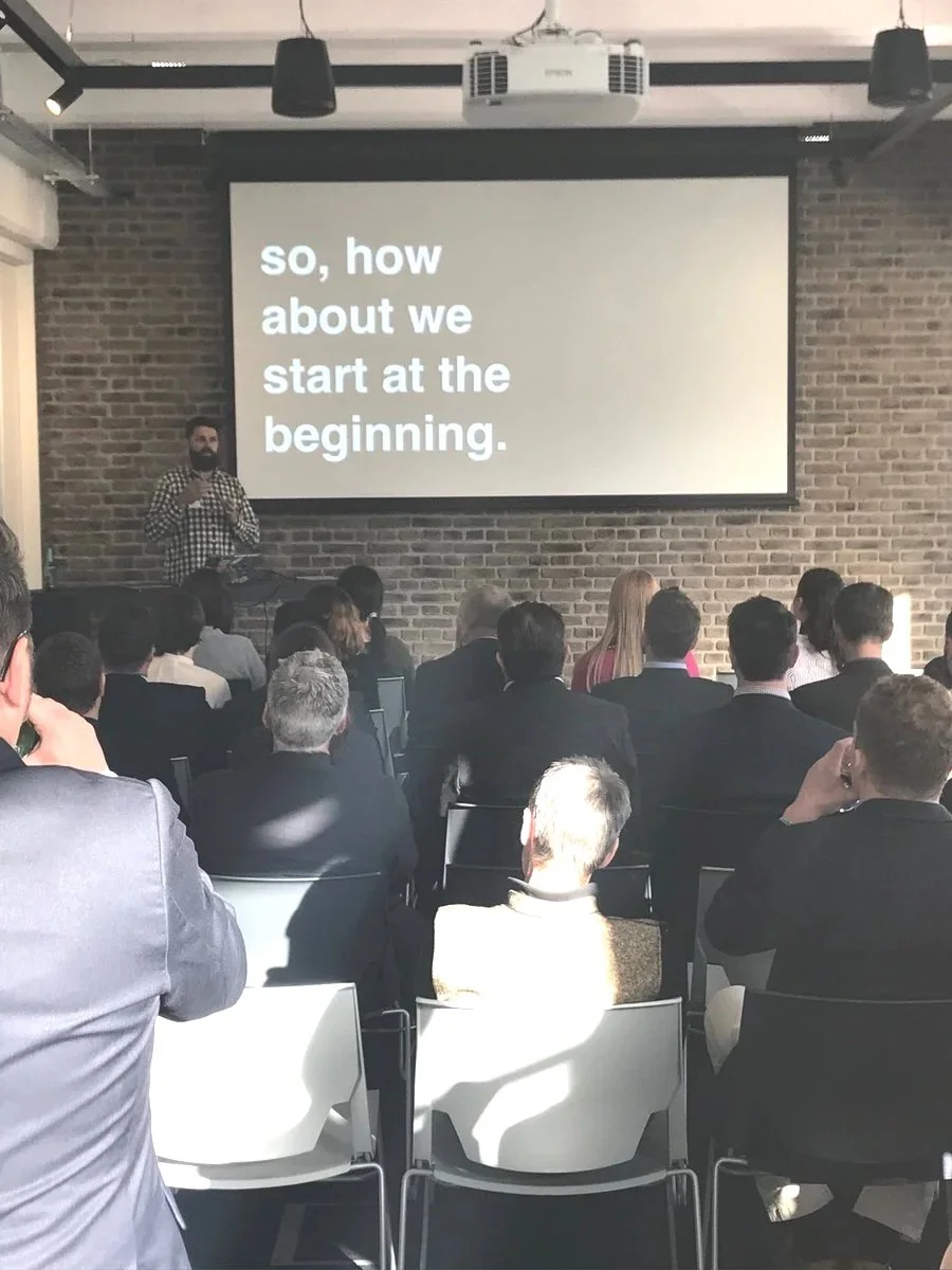 A presentation in a conference room with a speaker standing by a large screen that says, "so, how about we start at the beginning." The audience is seated, facing the speaker and screen, in a modern interior with exposed brick walls.