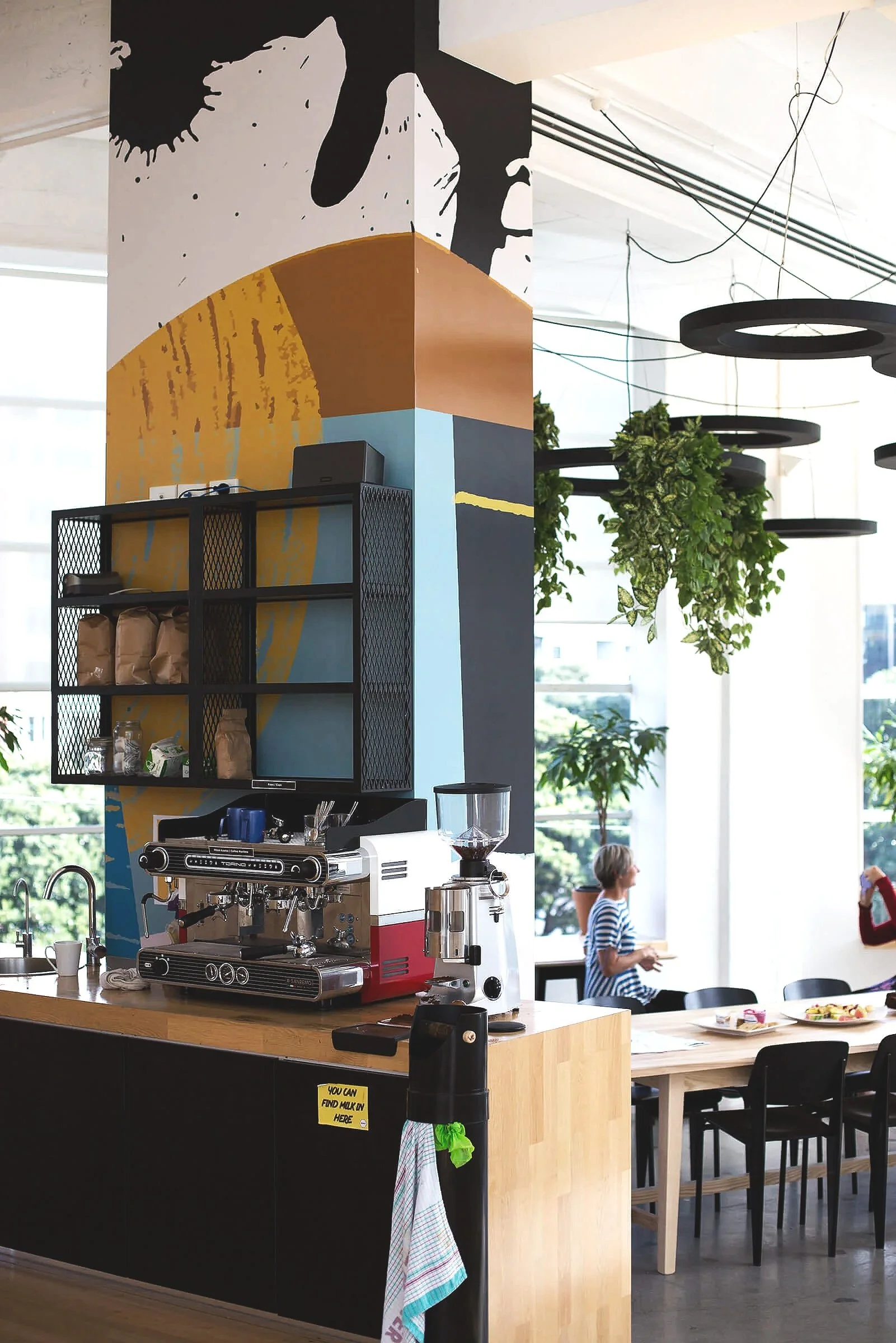 Interior of a modern cafe or restaurant with a coffee machine on a wooden counter, shelves with coffee bags, two people seated at a table, and hanging green plants in front of large windows.