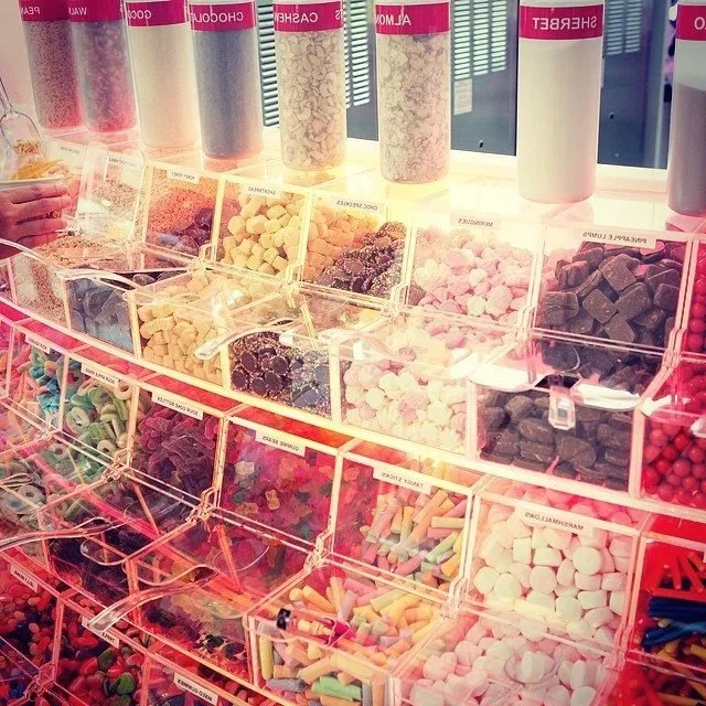 Display of assorted gummy candies in clear plastic bins and candy jars in a store, with pink-topped containers containing various candies placed above.