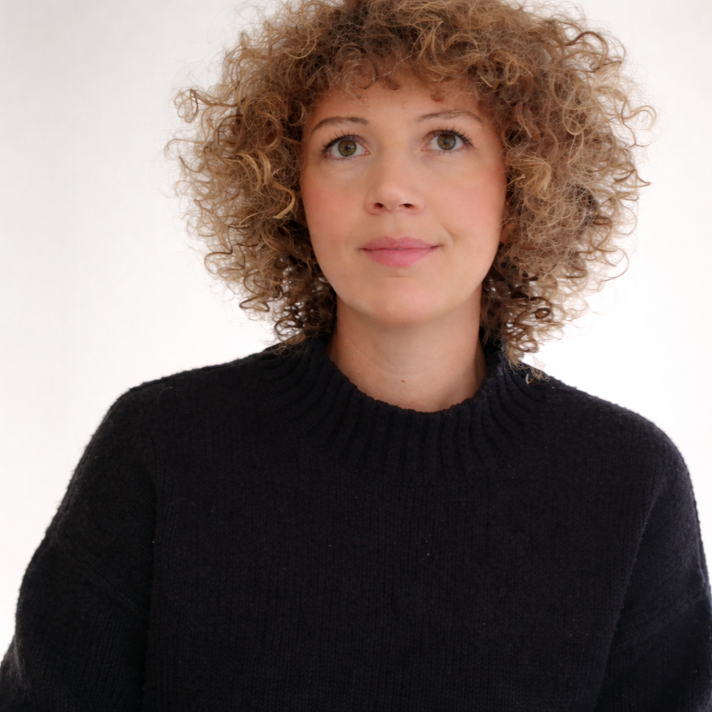 A woman with light skin and curly hair wearing a black sweater looks slightly upward with a neutral expression against a plain white background.