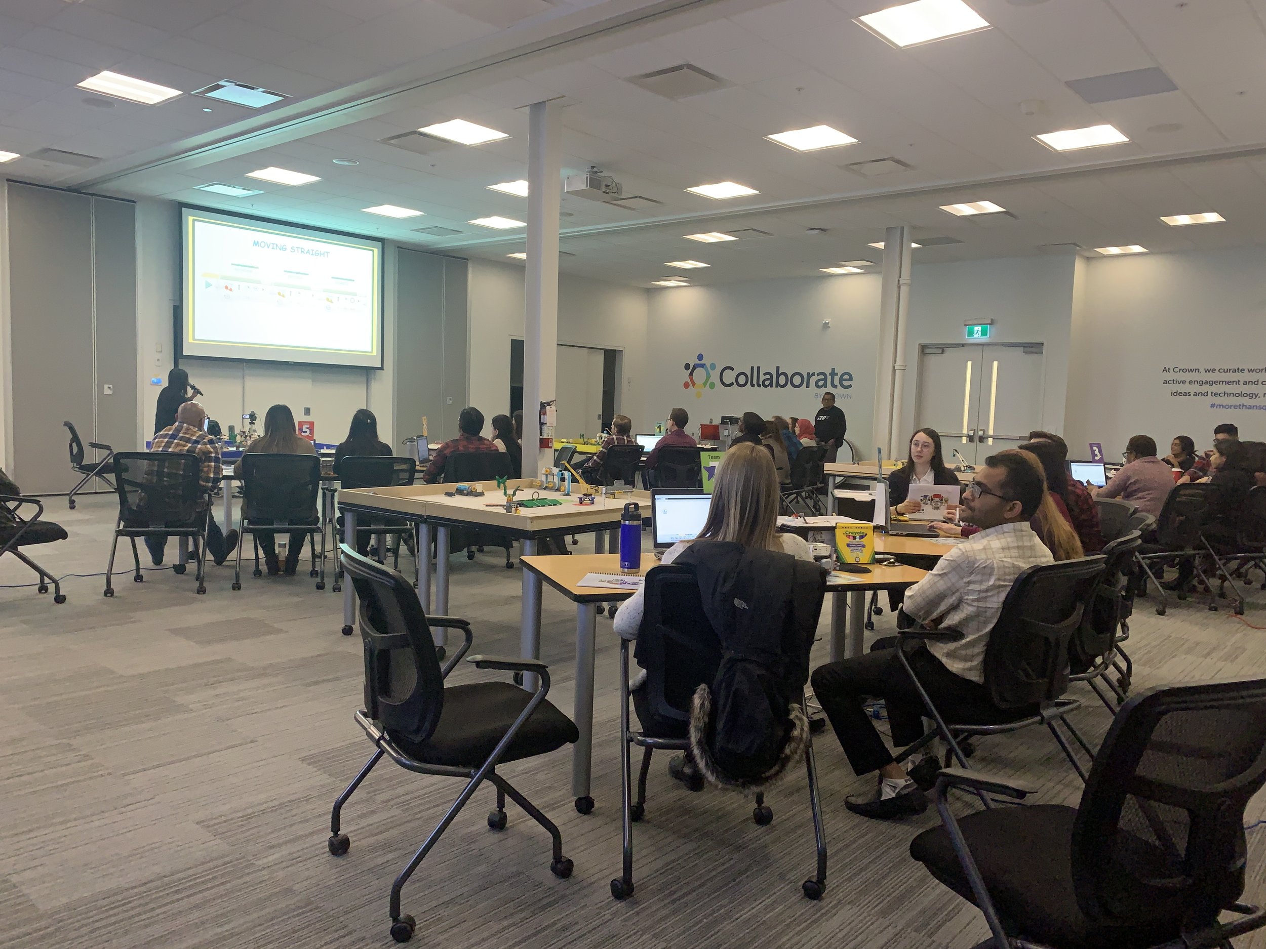 People attending a presentation in a conference room, with some working on laptops at tables, and a large screen displaying slides at the front. The room has a modern design with a sign that says 'Collaborate' on the wall.