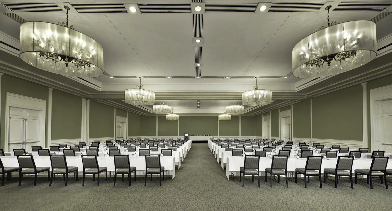 Large conference room with long tables arranged in a U-shape, black chairs with white cushions, white tablecloths, and large overhead chandeliers with crystal accents.