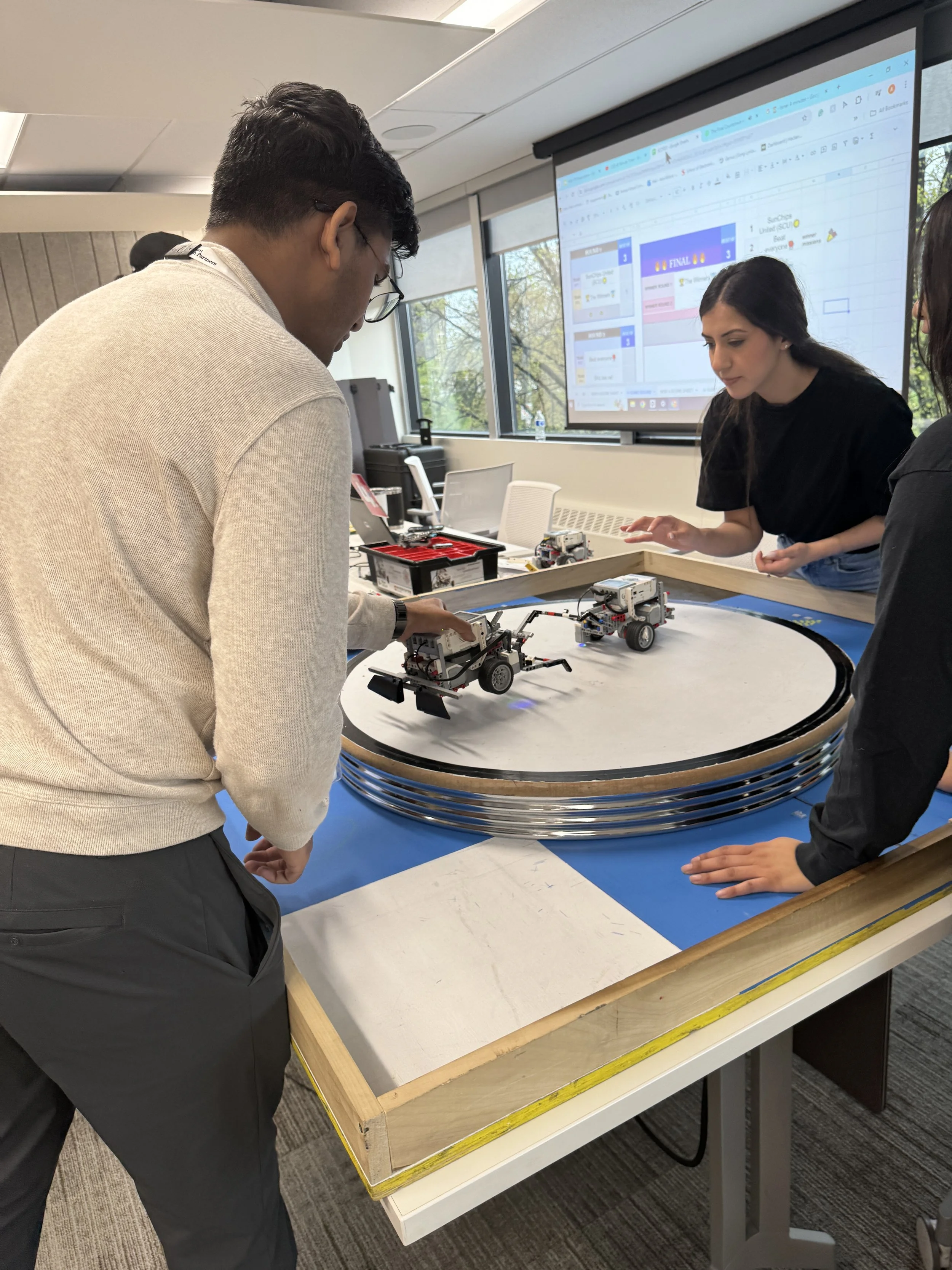 Students working on robot projects on a table with a circular track, while a large screen displays a digital scoreboard or project interface in the background.