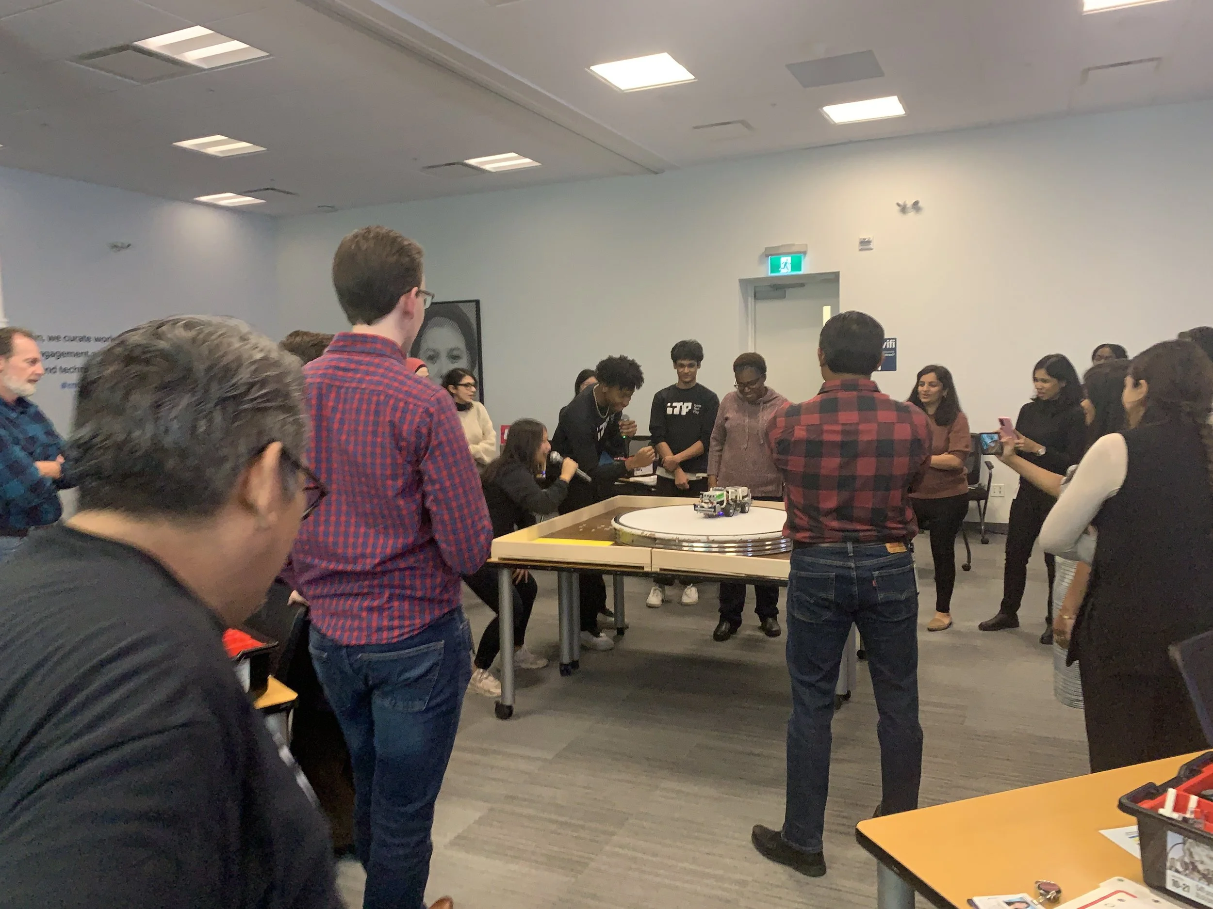 People gathered in a room watching a robot on a circular track, some taking photos or videos, with a diverse group including young adults and others.