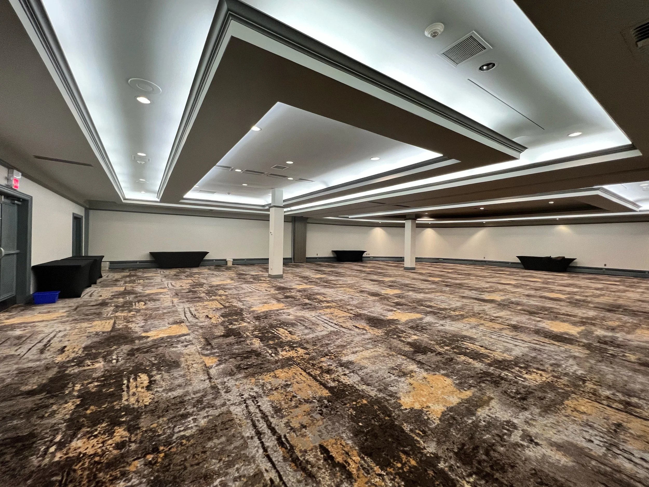 Large empty conference room with patterned carpet, white walls, and a decorative ceiling with recessed lighting, three black tables along the walls, and a few white columns.