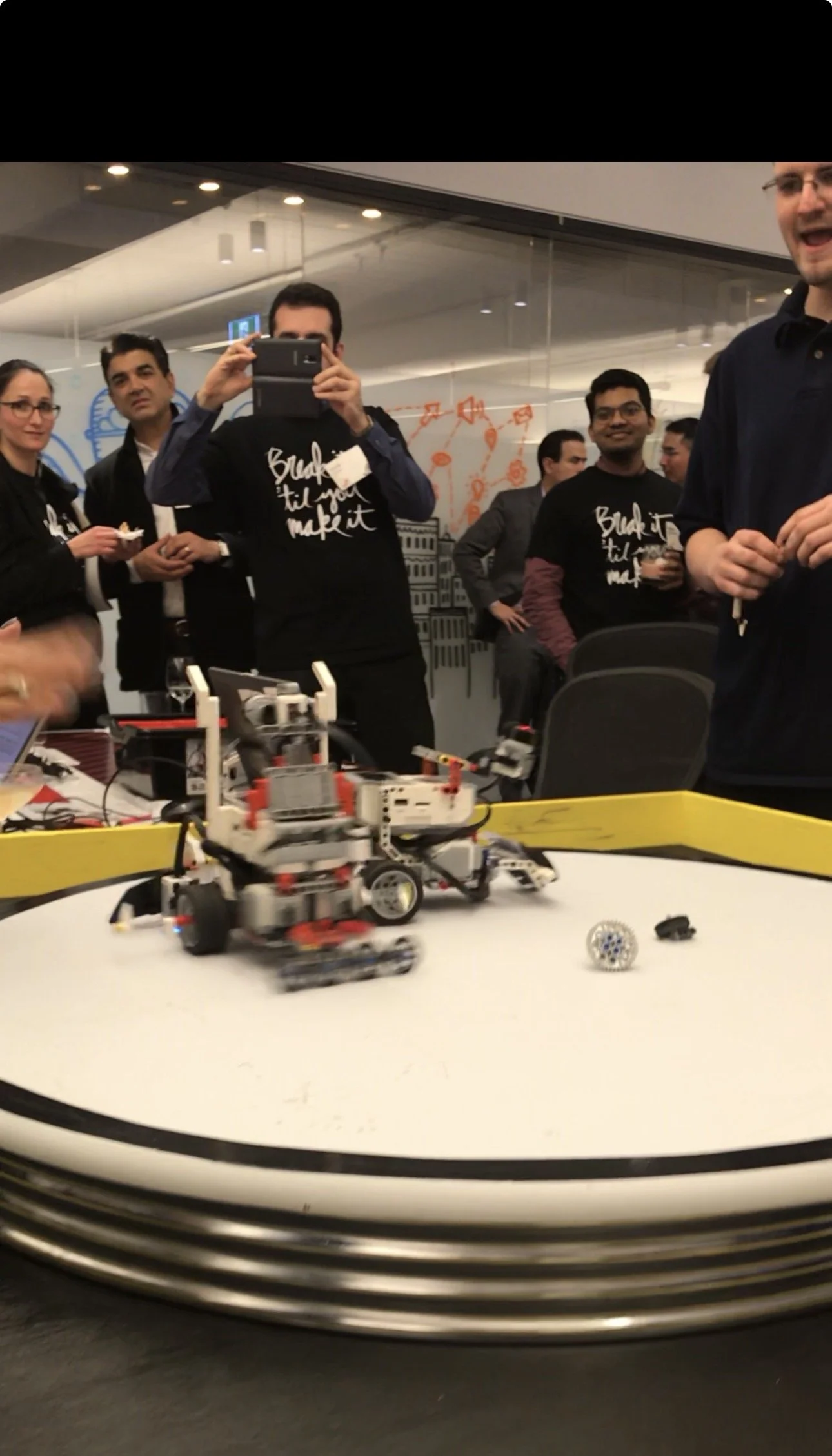 Group of people gathered around a robotics competition table, with a robot in motion on the table, while some individuals wear matching black t-shirts that say "Build it till you make it." One person is taking a photo with a mobile device.