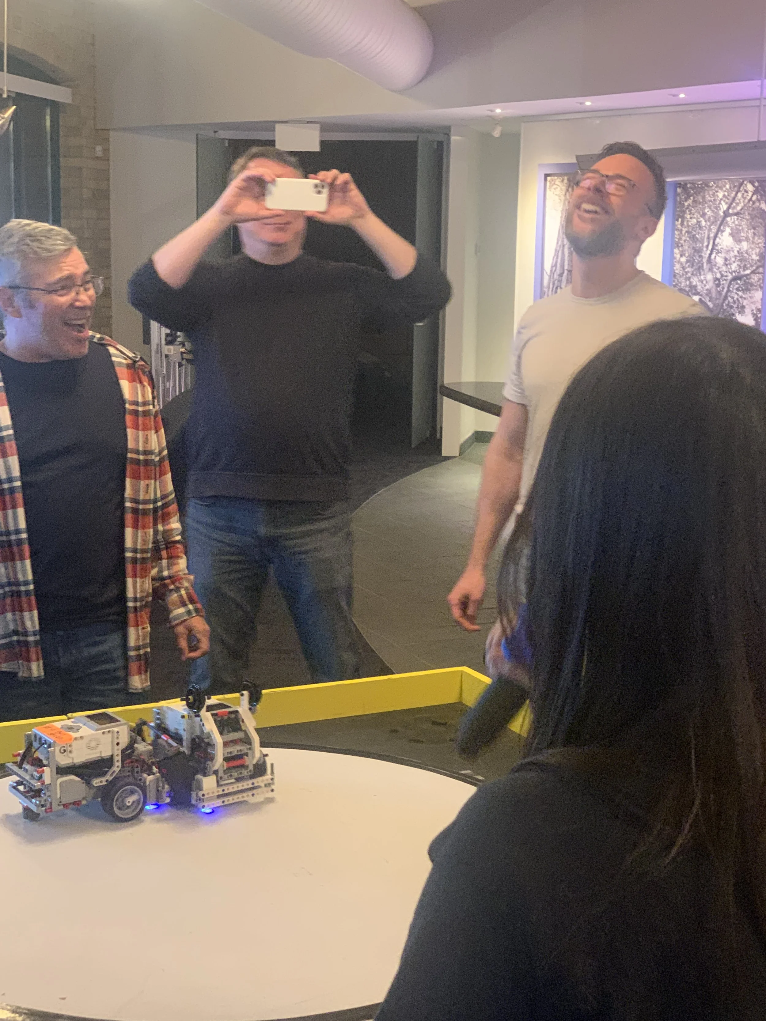 Group of people gathered around a table with a small robot, laughing and taking pictures at a robotics event or team activity.