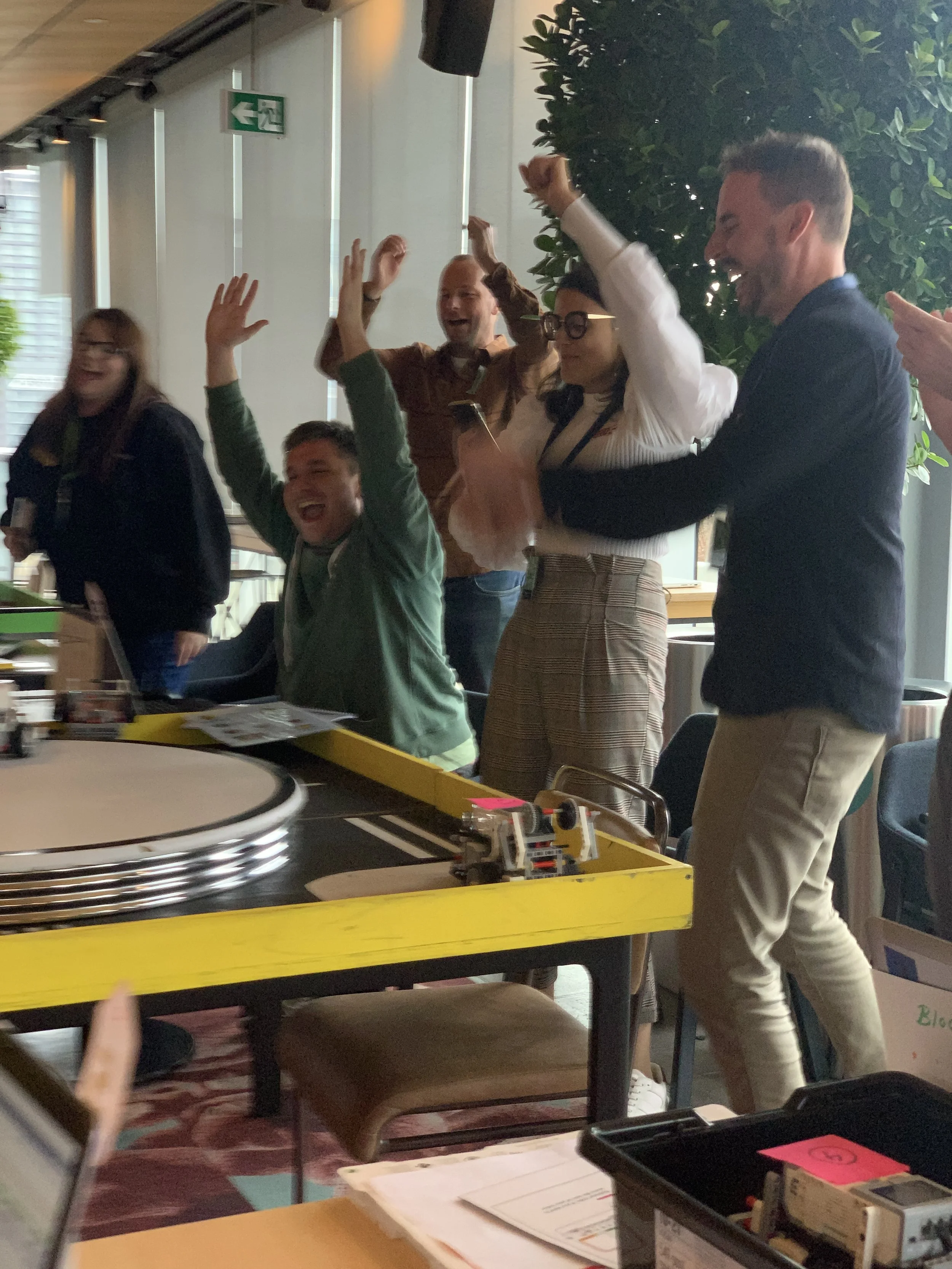 Group of five people in an office celebrating, some with hands raised, smiling and laughing, around a table with a yellow border, a LEGO setup, and documents.
