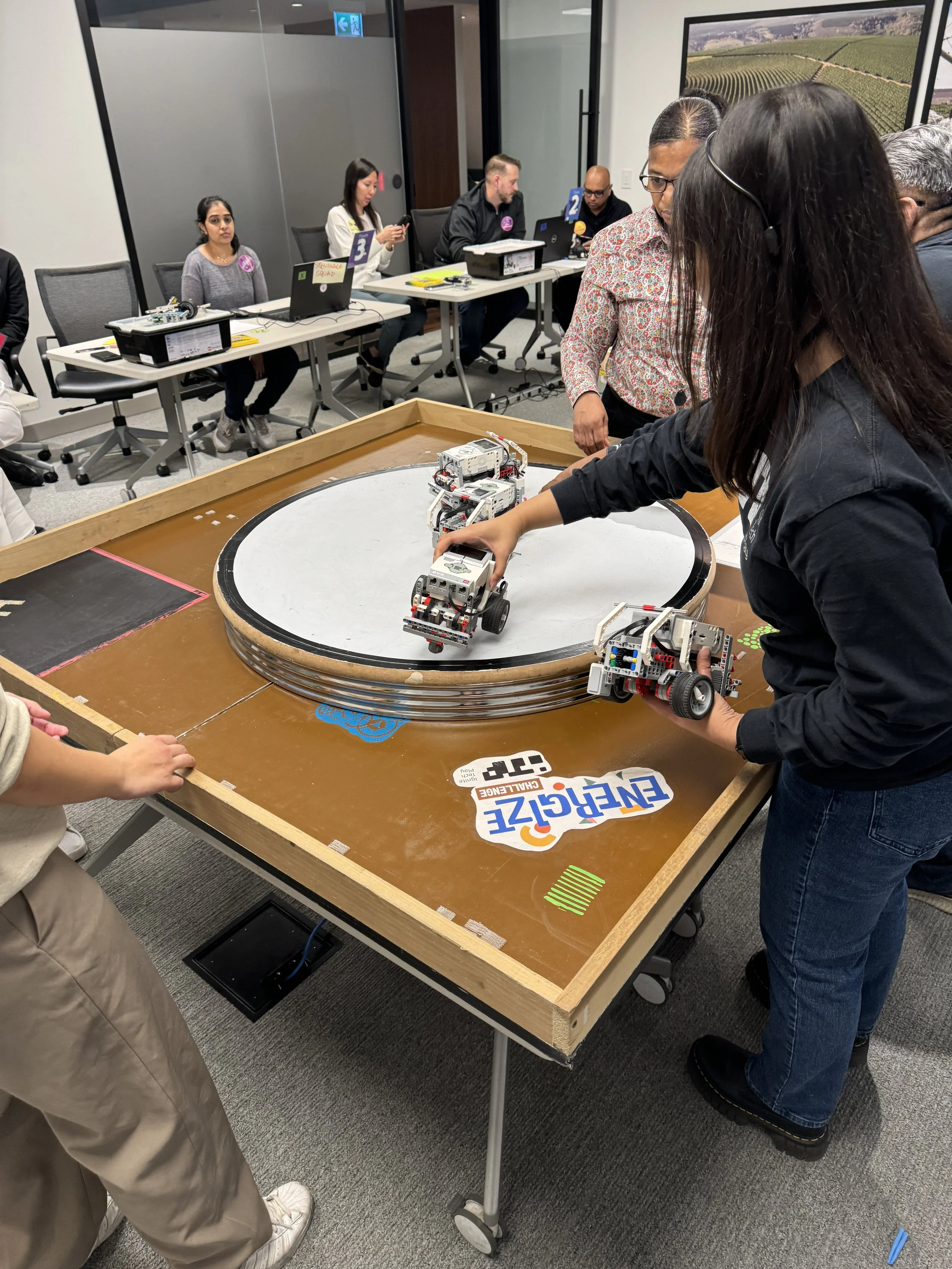 People operating and observing small robots on a circular track in a classroom or conference room setting, with others working on laptops at tables in the background.