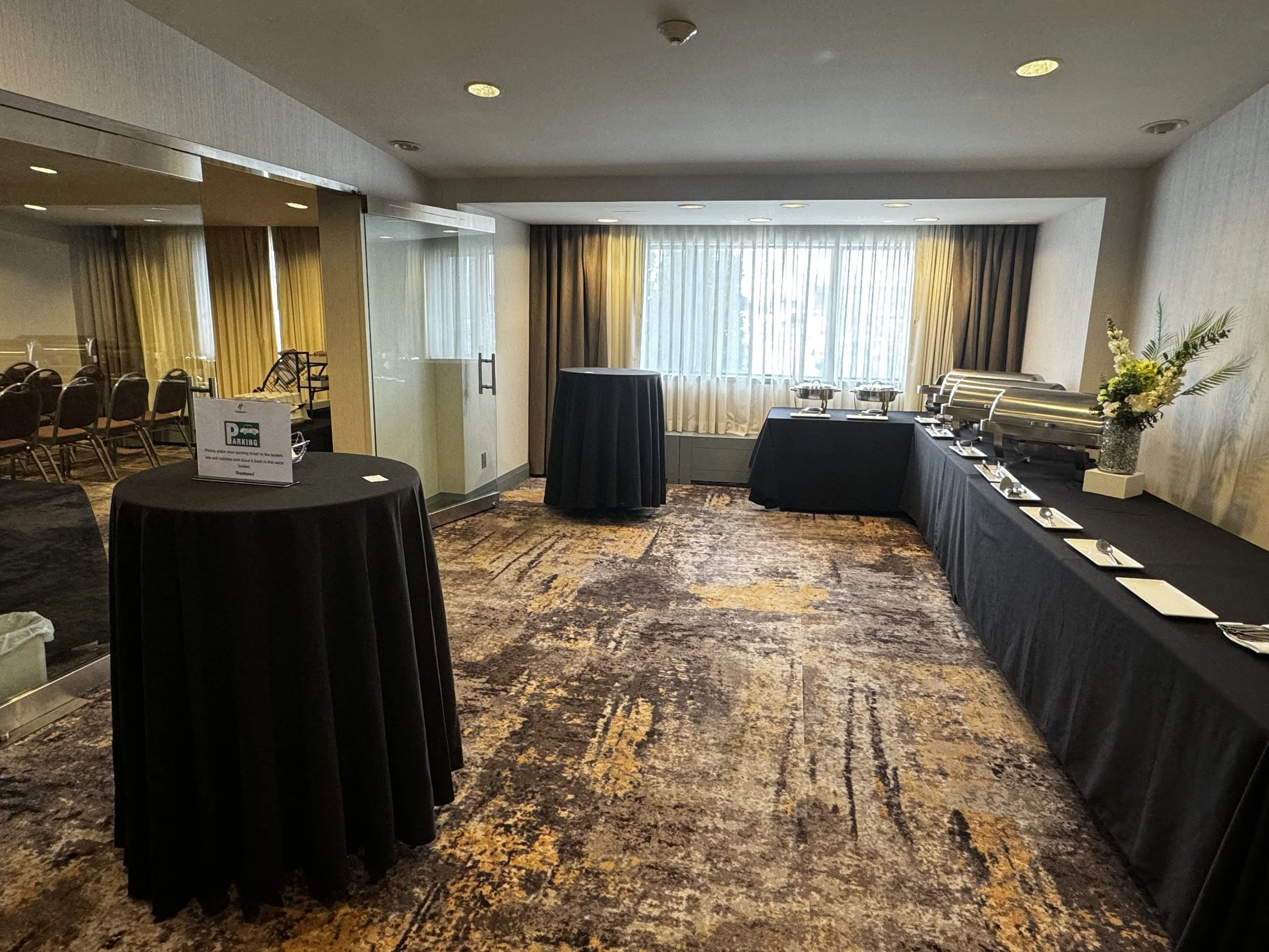 Empty banquet or conference room with buffet table, high top table, window with curtains, and chairs visible through glass door.