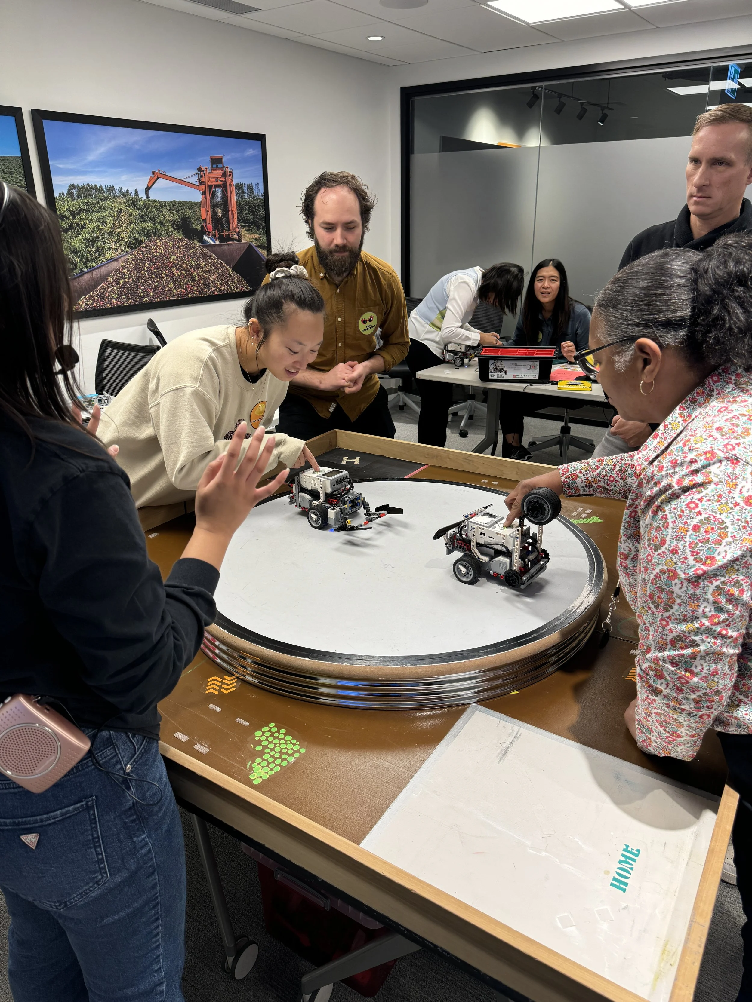 Group of people gathered around a circular robotics table, with two robots in motion on the table, in a room with posters of agricultural machinery on the wall.