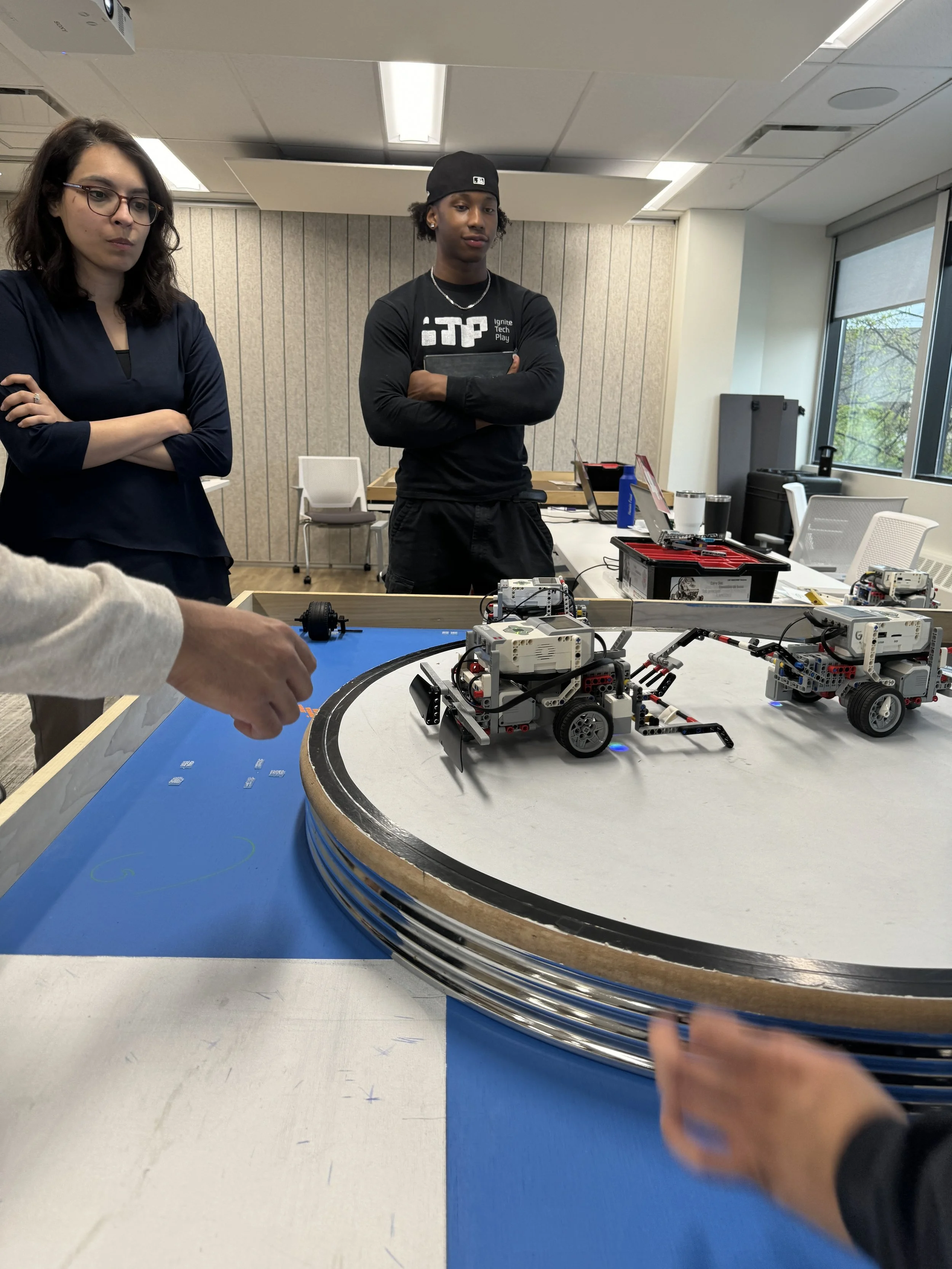 People observing a robotics demonstration with small robotic vehicles on a track surface, in a well-lit room with windows and equipment in the background.