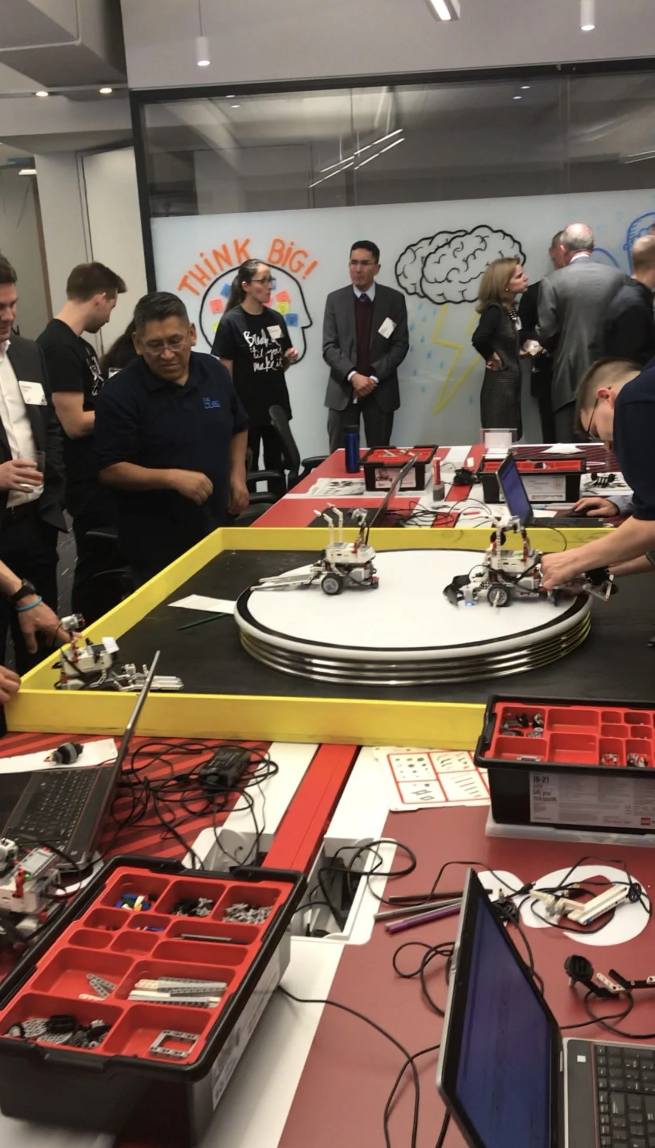 People gathered around a table with robotics kits, working on robot projects, in a room with a large window and colorful drawings on the wall.