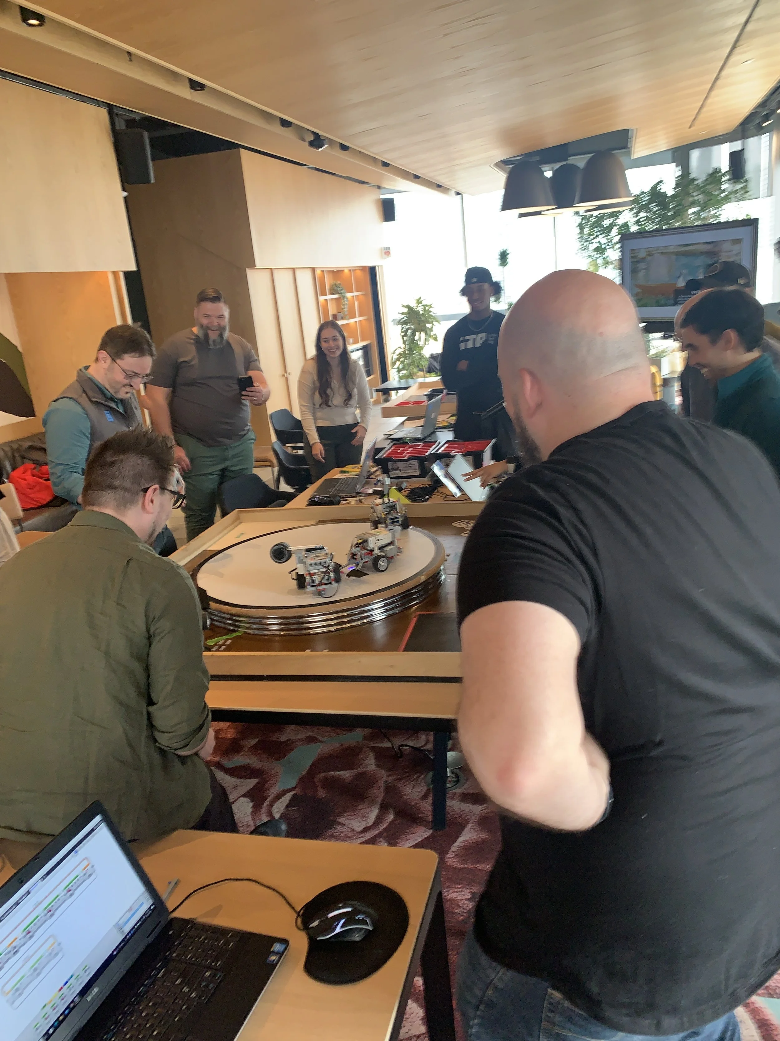 A group of people gathered around a table with a robotic race track, playing with small robots during an event or workshop in a modern conference room.