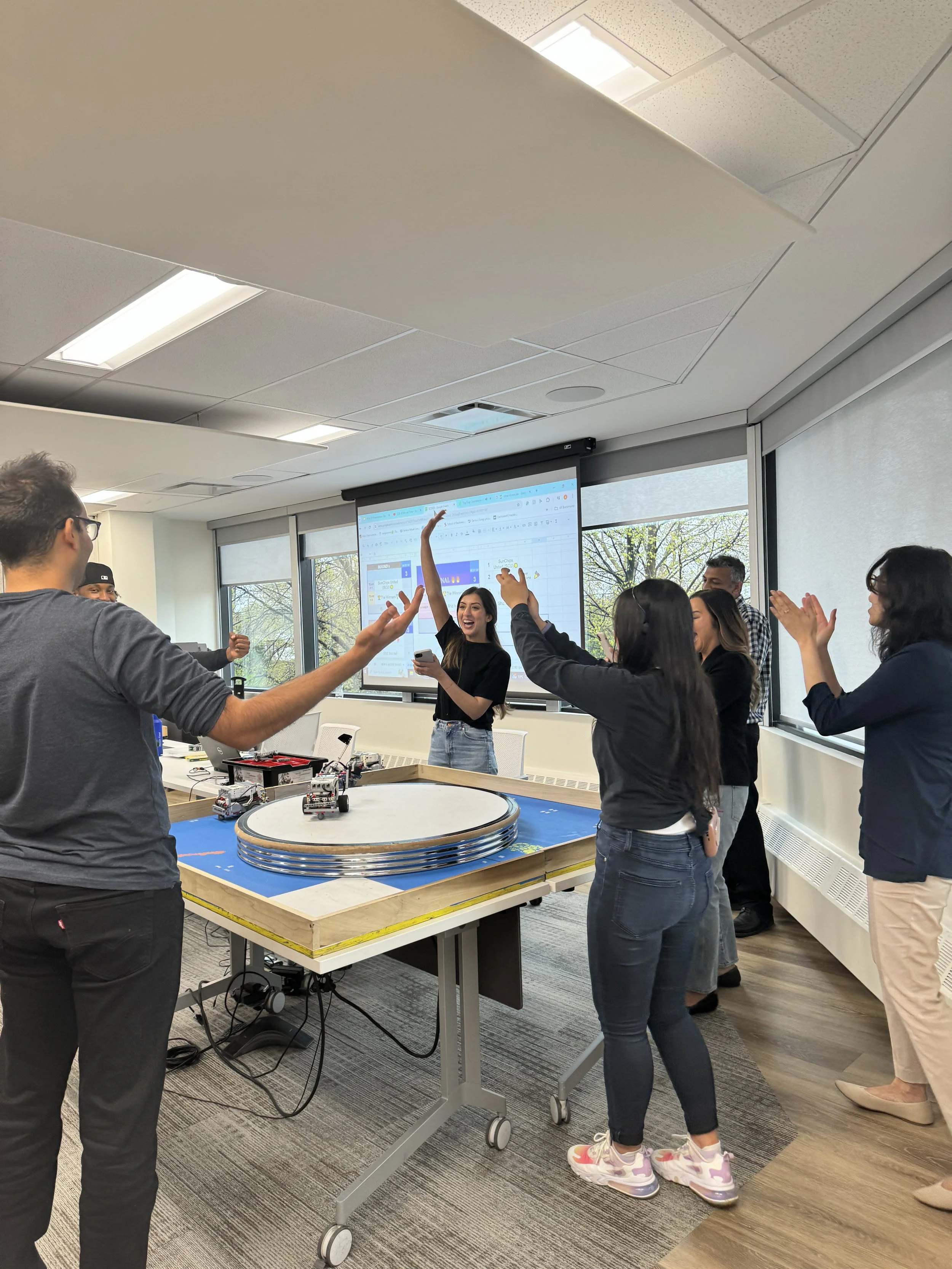 Group of people celebrating around a robotics competition table in a conference room with large windows and a projected presentation