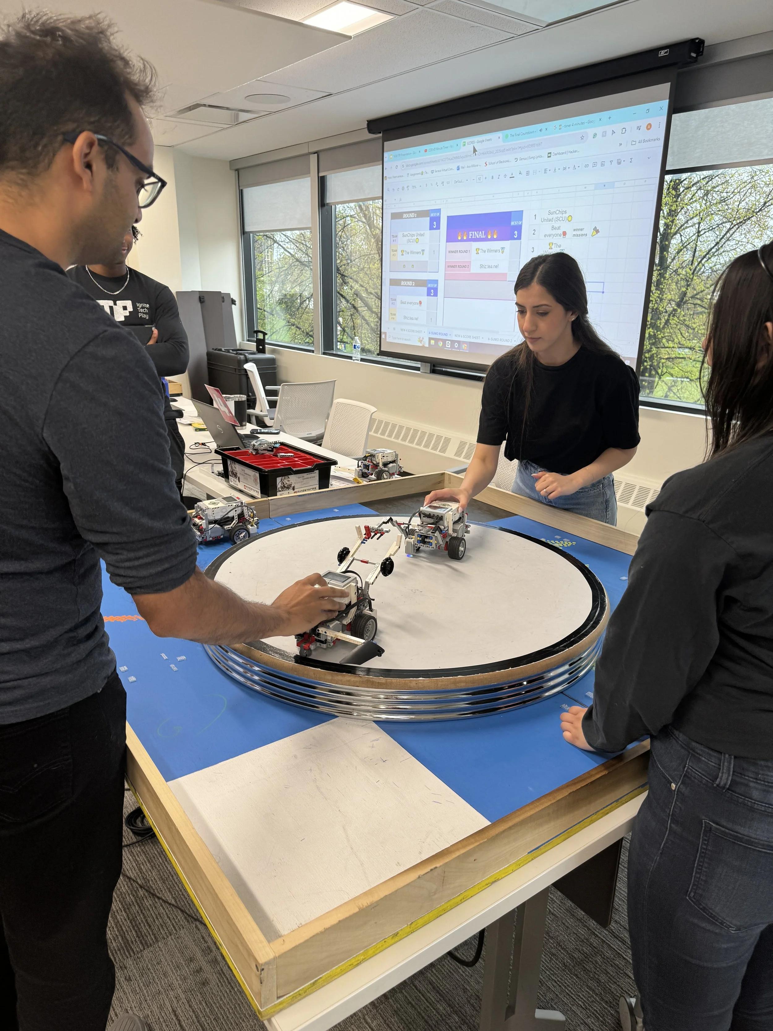 People working with LEGO Mindstorm robots on a circular robot arena at a workshop or classroom.