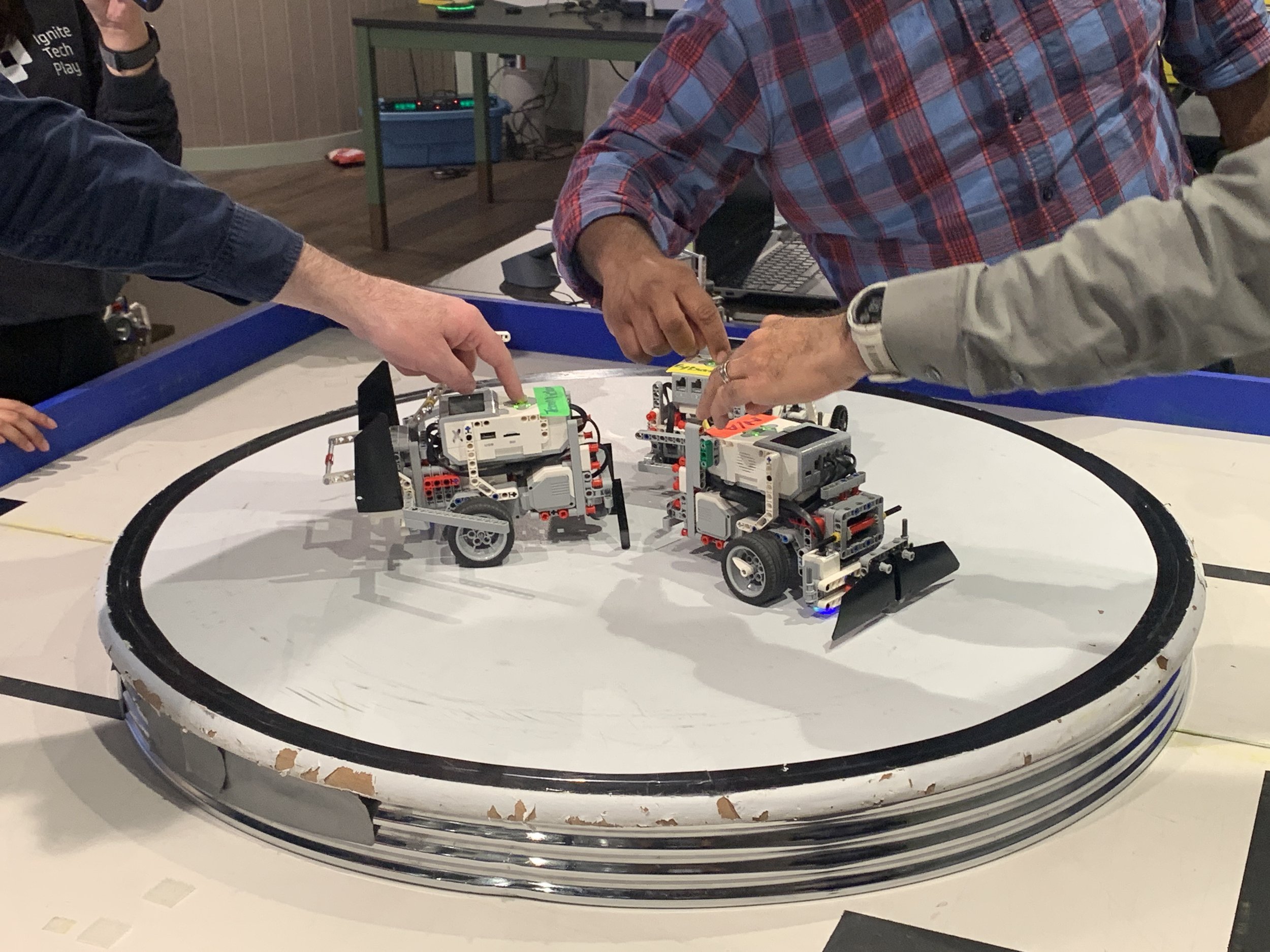 Multiple people are working on robots on a circular platform, inside a designated area marked by a black border. The robots are small, wheeled, and have various electronic components.