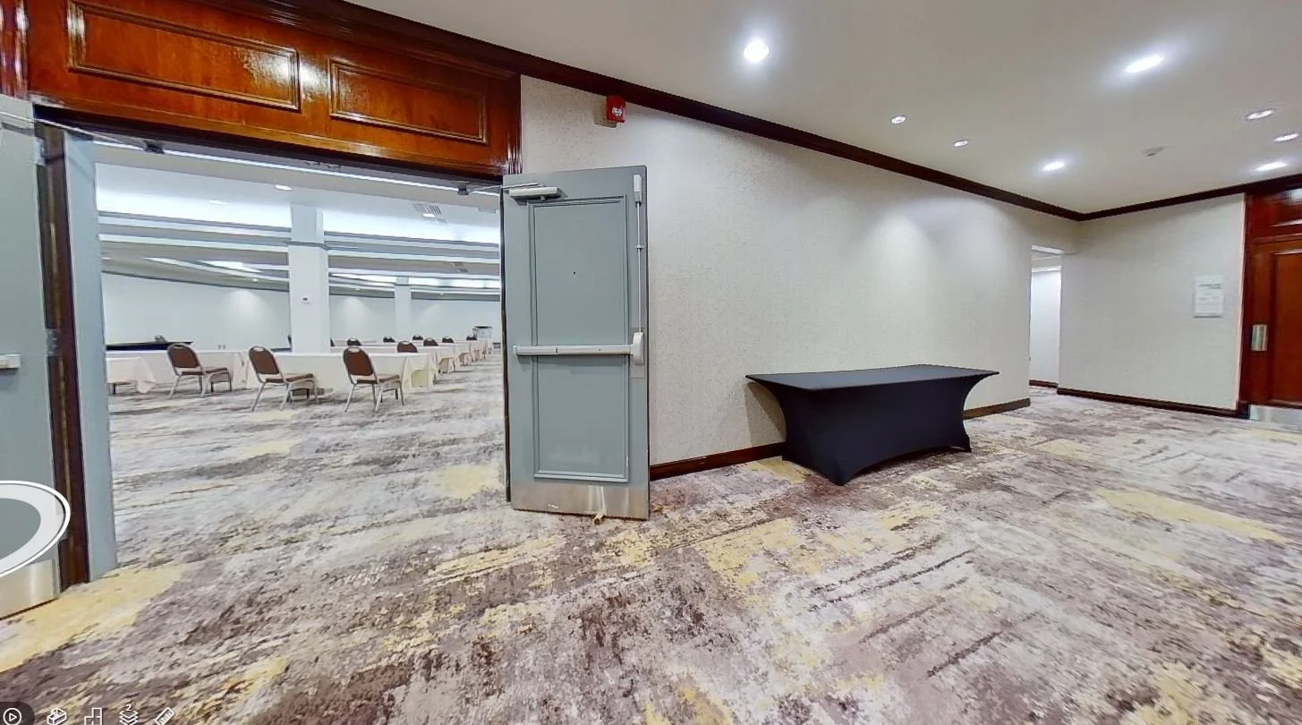 Empty conference room with round tables and chairs, viewed through a doorway with dark wood trim; carpeted floor with abstract pattern, and plain white walls with a fire alarm on one wall.
