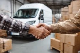 Two people shaking hands in a warehouse with a truck and boxes in the background.