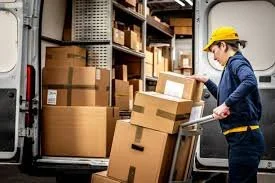 A woman wearing a yellow safety helmet loading boxes onto a cart outside a delivery van in a warehouse.
