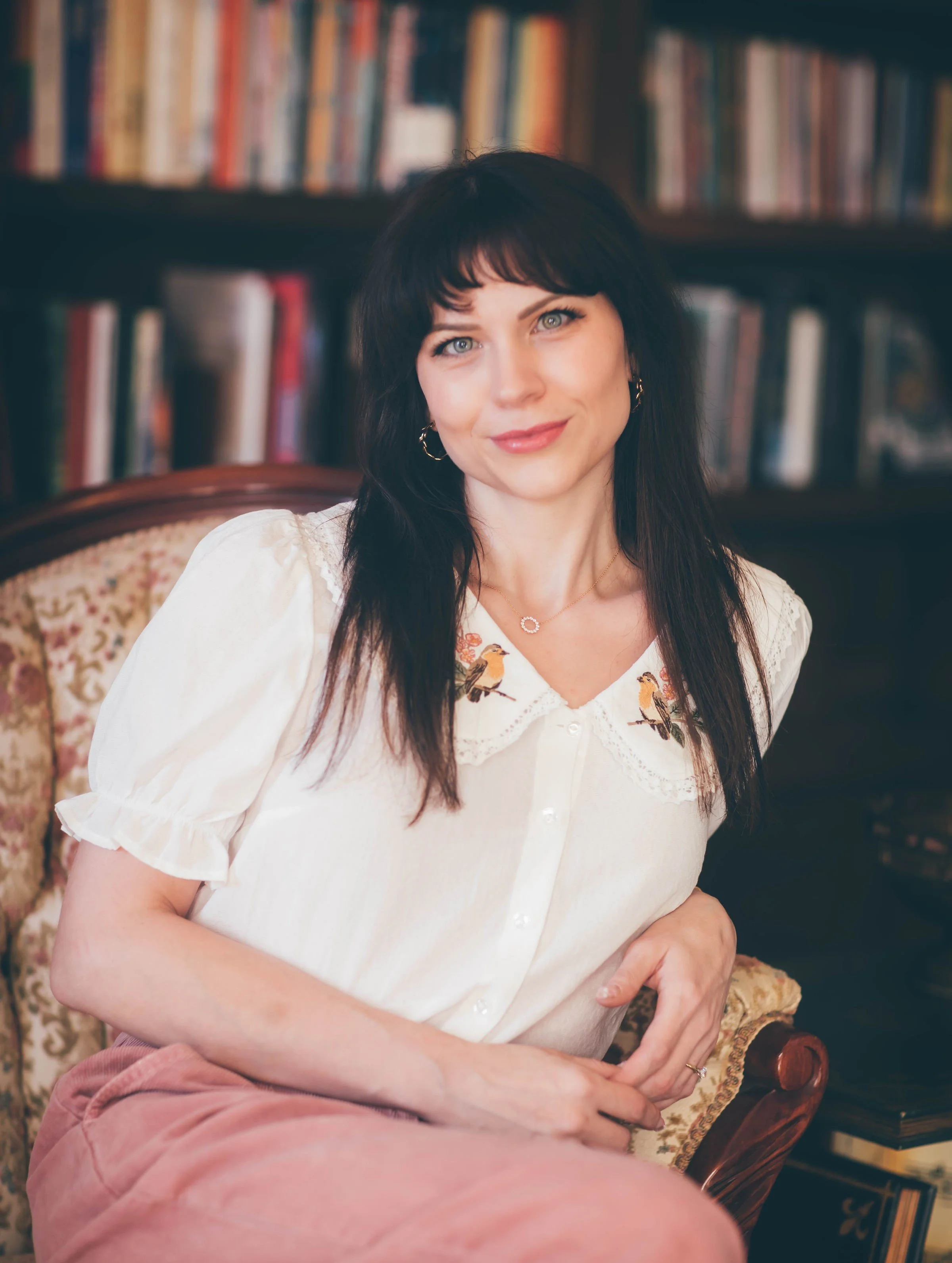 Oval portrait of poet Chrissy Martin seated in a patterned armchair, wearing a white blouse and pink skirt, with dark hair and bangs. She faces the camera with a soft smile; blurred bookshelves fill the background.