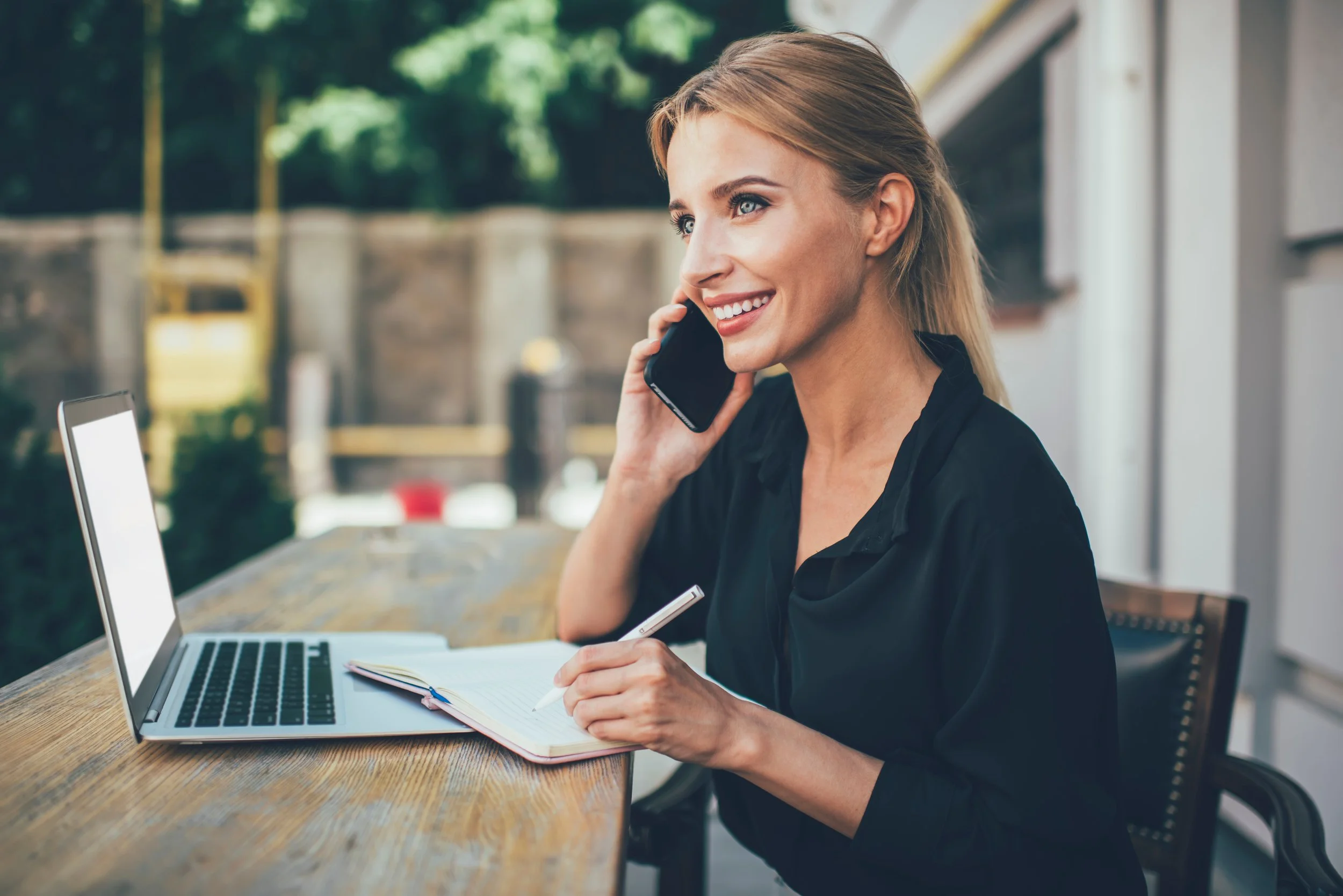 Woman sitting at outdoor table with laptop, notebook, and pen, talking on cell phone, smiling
