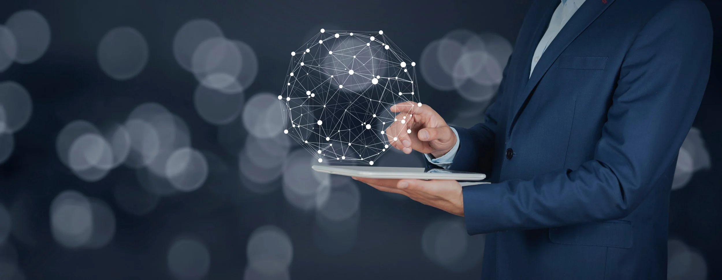 Businessperson in a navy suit holding a tablet with a digital globe made of interconnected dots and lines floating above it, against a background of blurred lights.