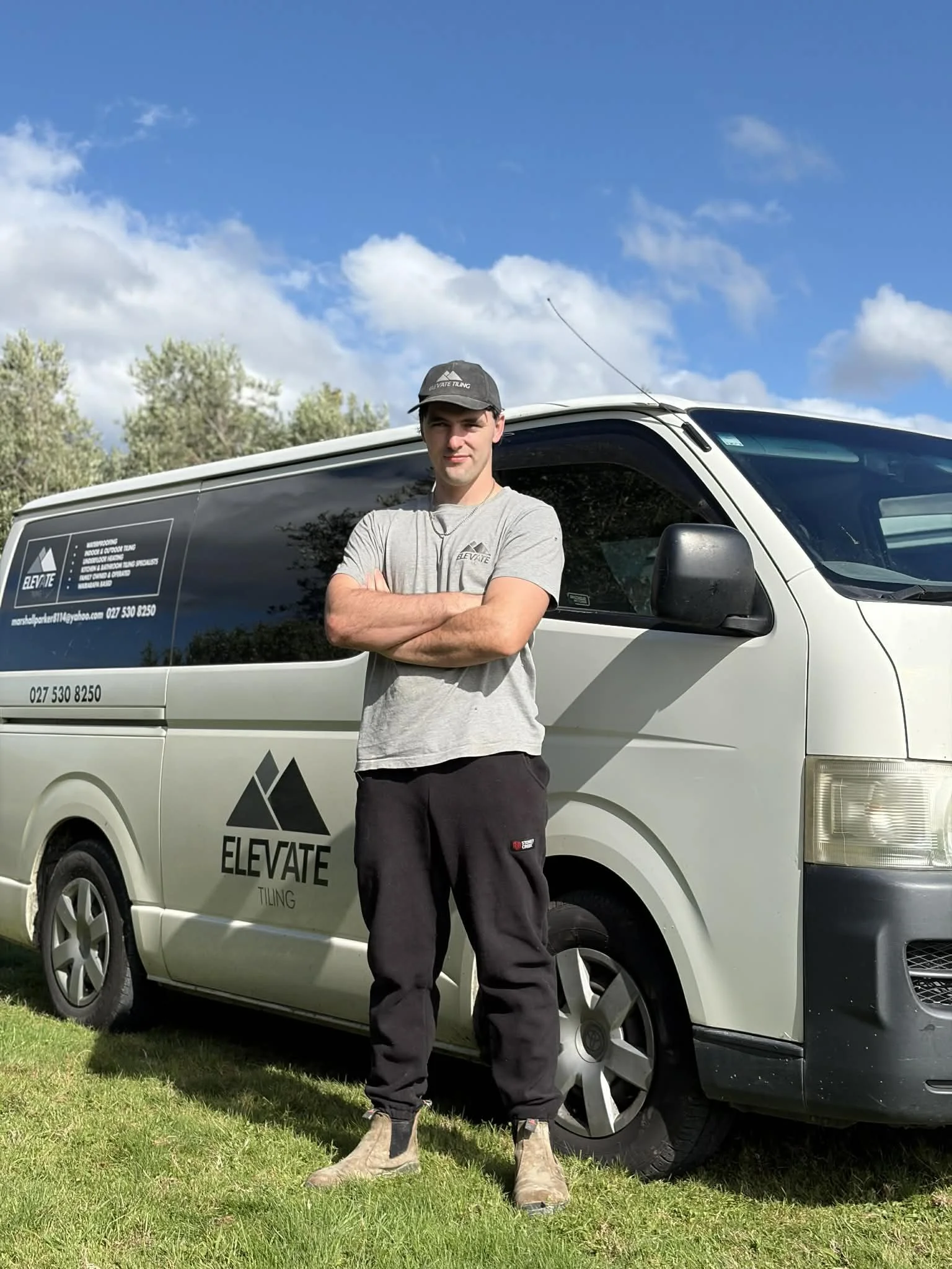 A woman with curly dark hair sitting on a bench in a sunlit room, wearing a sleeveless beige top and loose beige trousers, looking thoughtfully into the distance.