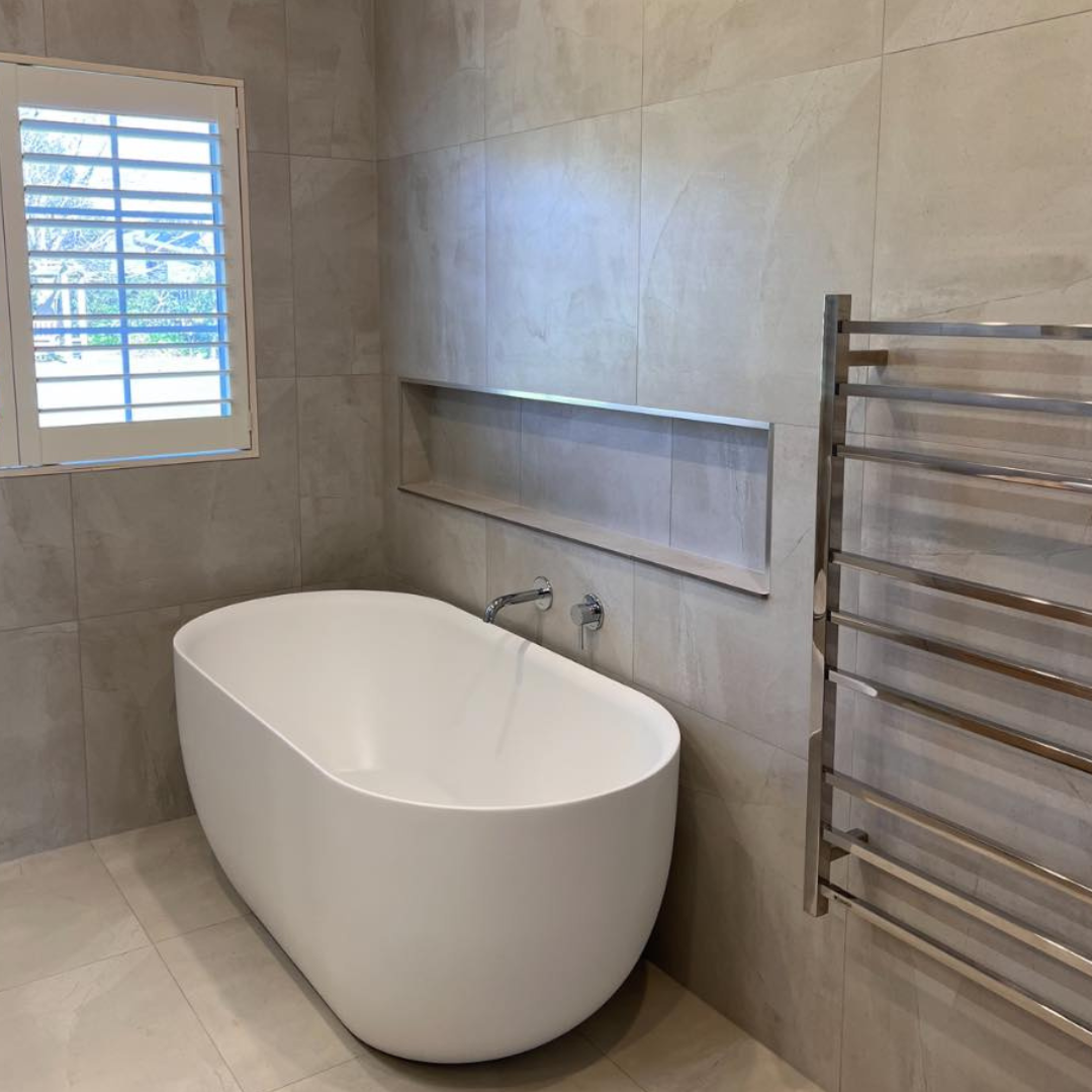 Modern bathroom with a standalone oval bathtub, a window with shutters, a built-in wall niche, and a stainless steel towel warmer, with beige tiled walls and floor.