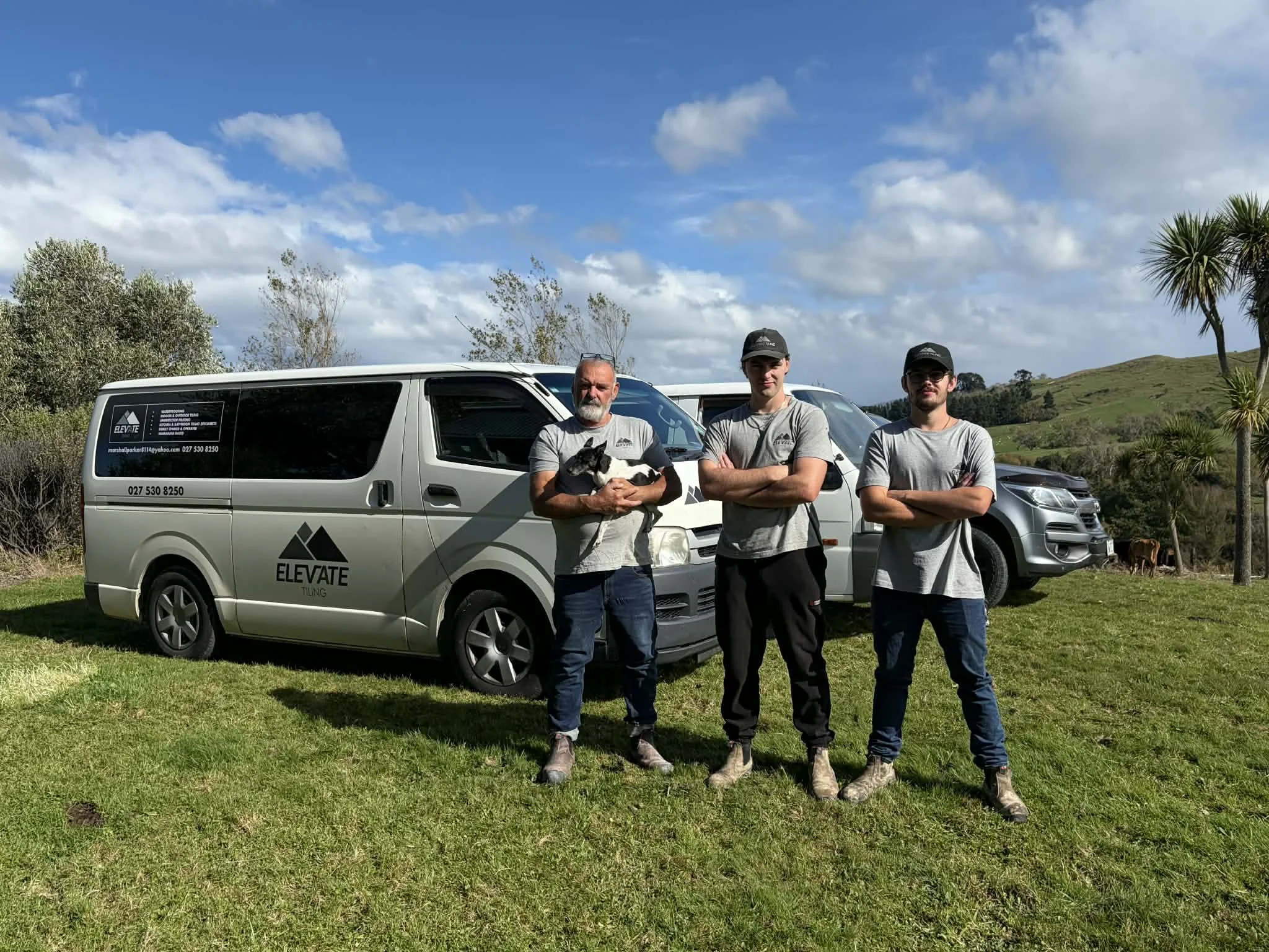 Three men in gray shirts and work boots standing on grass in front of two company vans with 'Elevate Tiling' logo. One man is holding a black and white dog. A landscape with trees, hills, and a partly cloudy sky is visible in the background.