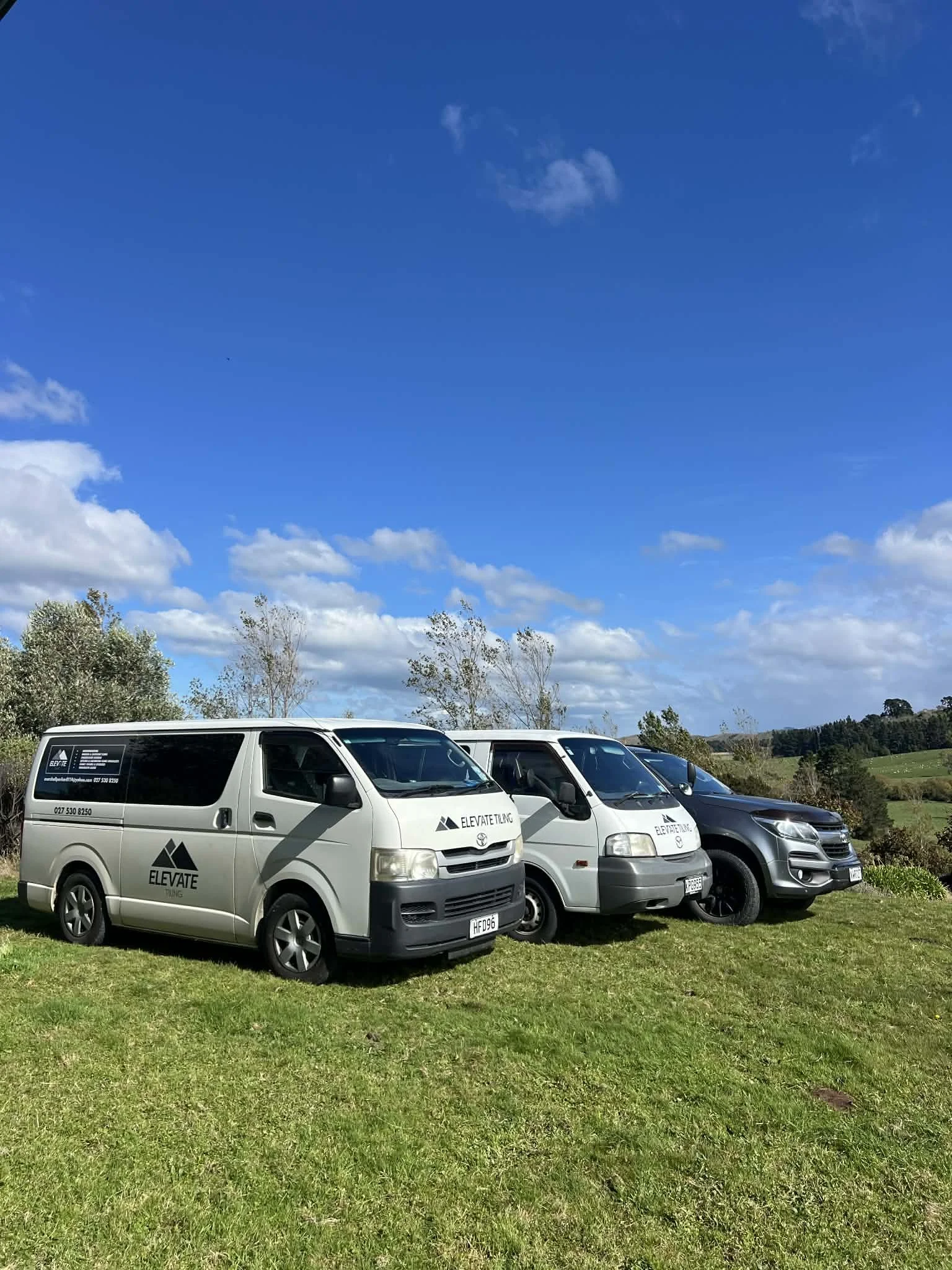 Three vehicles parked on a grassy field under a bright blue sky with scattered clouds. Two vans with 'Elevate Tiling' logos are on the left, and a black SUV is on the right.