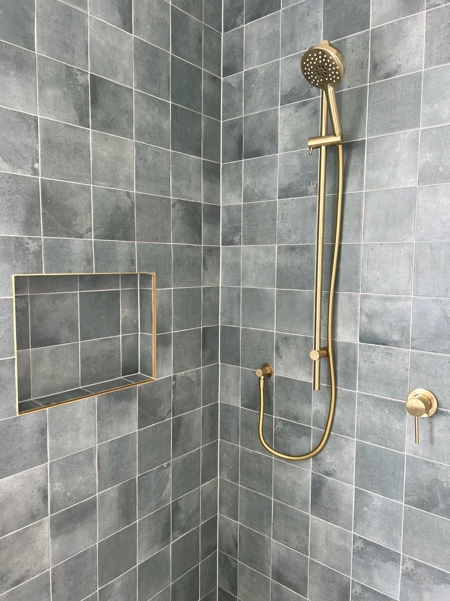A close-up view of a shower with gray tiled walls, a built-in square shelf with gold trim, a gold handheld showerhead, and a gold wall-mounted control knob.