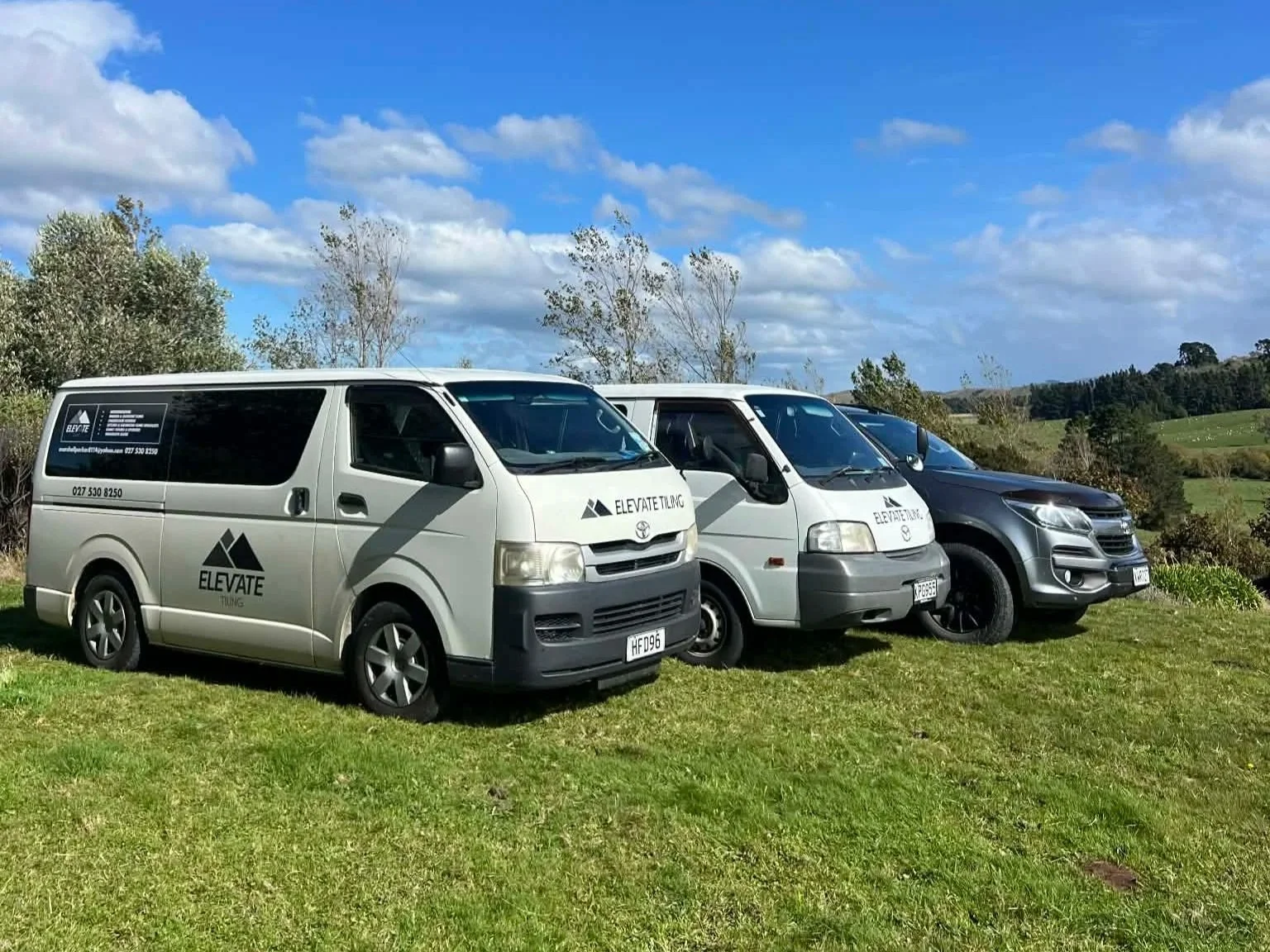 Three vans parked on a grassy field with a hilly rural landscape and partly cloudy blue sky in the background.