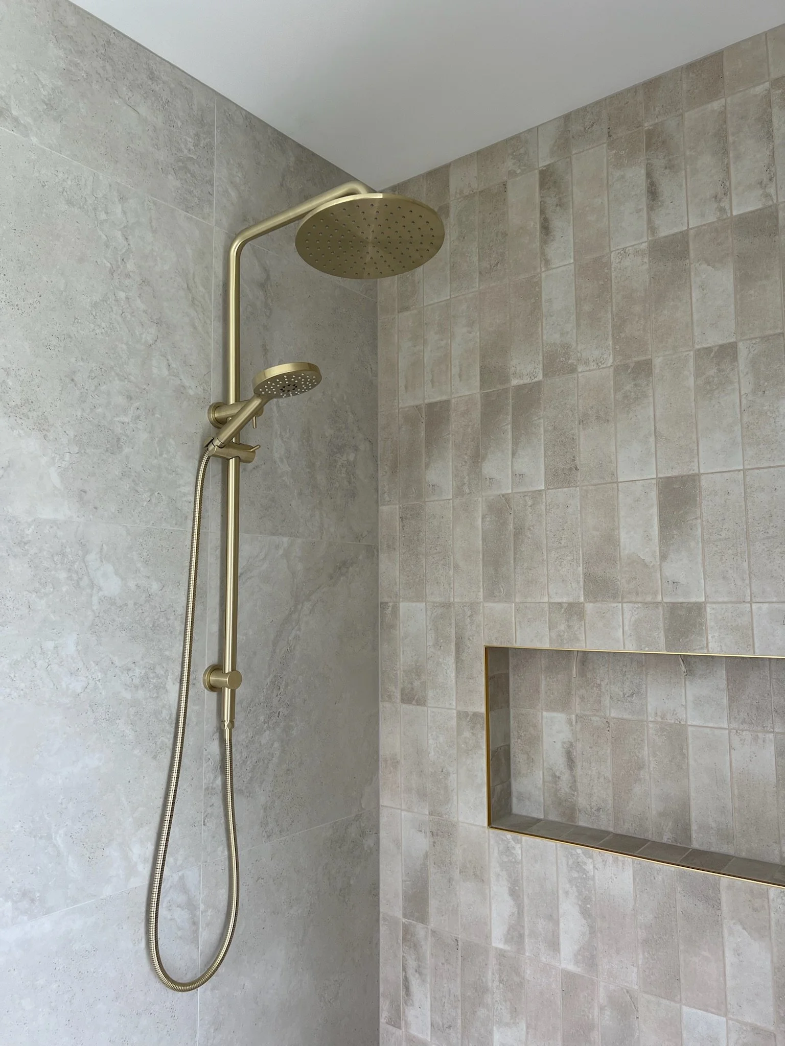 Shower area with a gold-colored rain showerhead and handheld shower wand mounted on a wall with beige tiles, including a built-in niche with a gold trim.