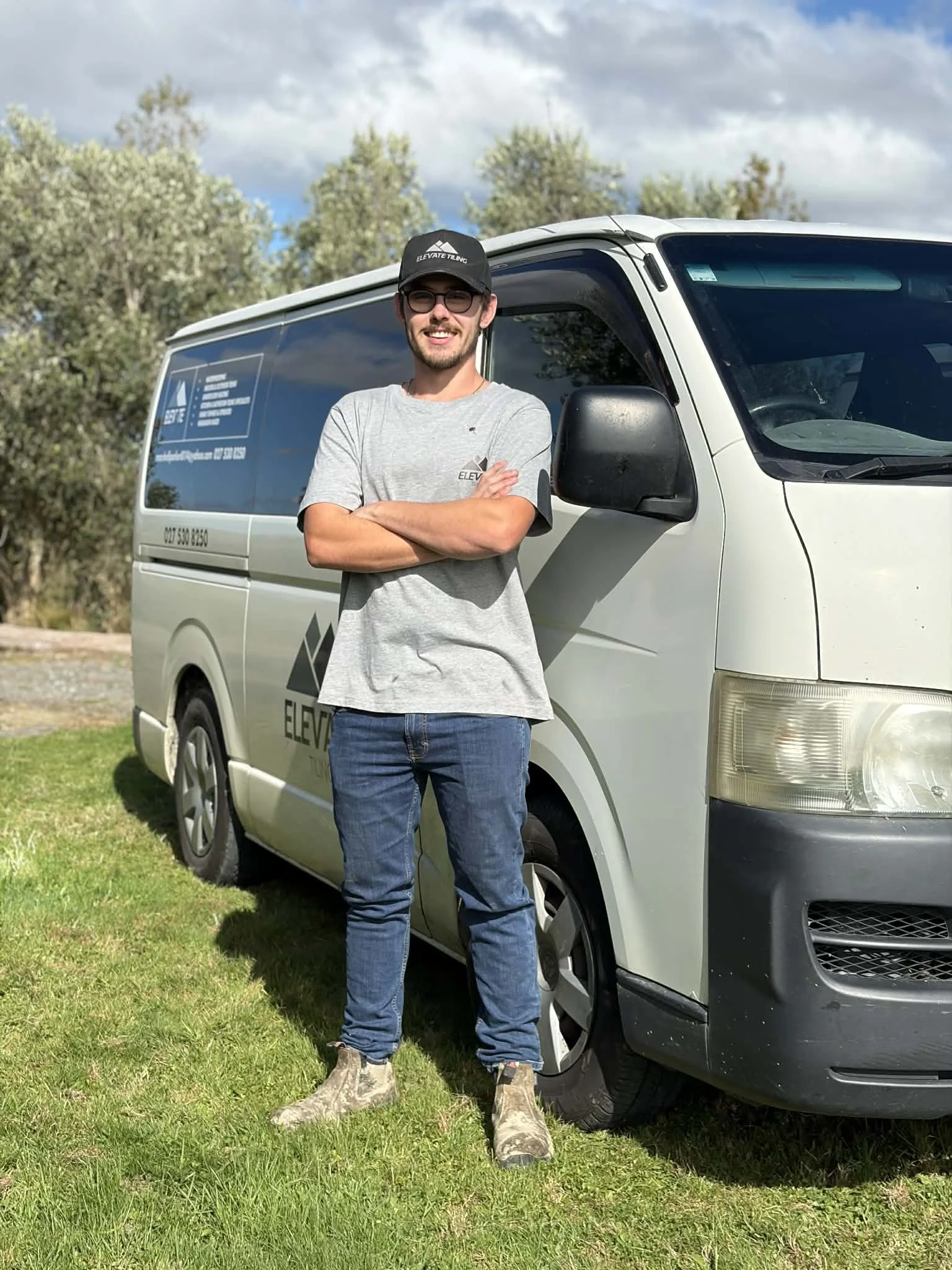 A woman with short blonde hair wearing an orange sleeveless top and beige pants, sitting on a beige bench, writing in a notebook. There is a large green plant on her left and a framed abstract art piece with earthy tones on her right, with sunlight casting shadows on the wall.