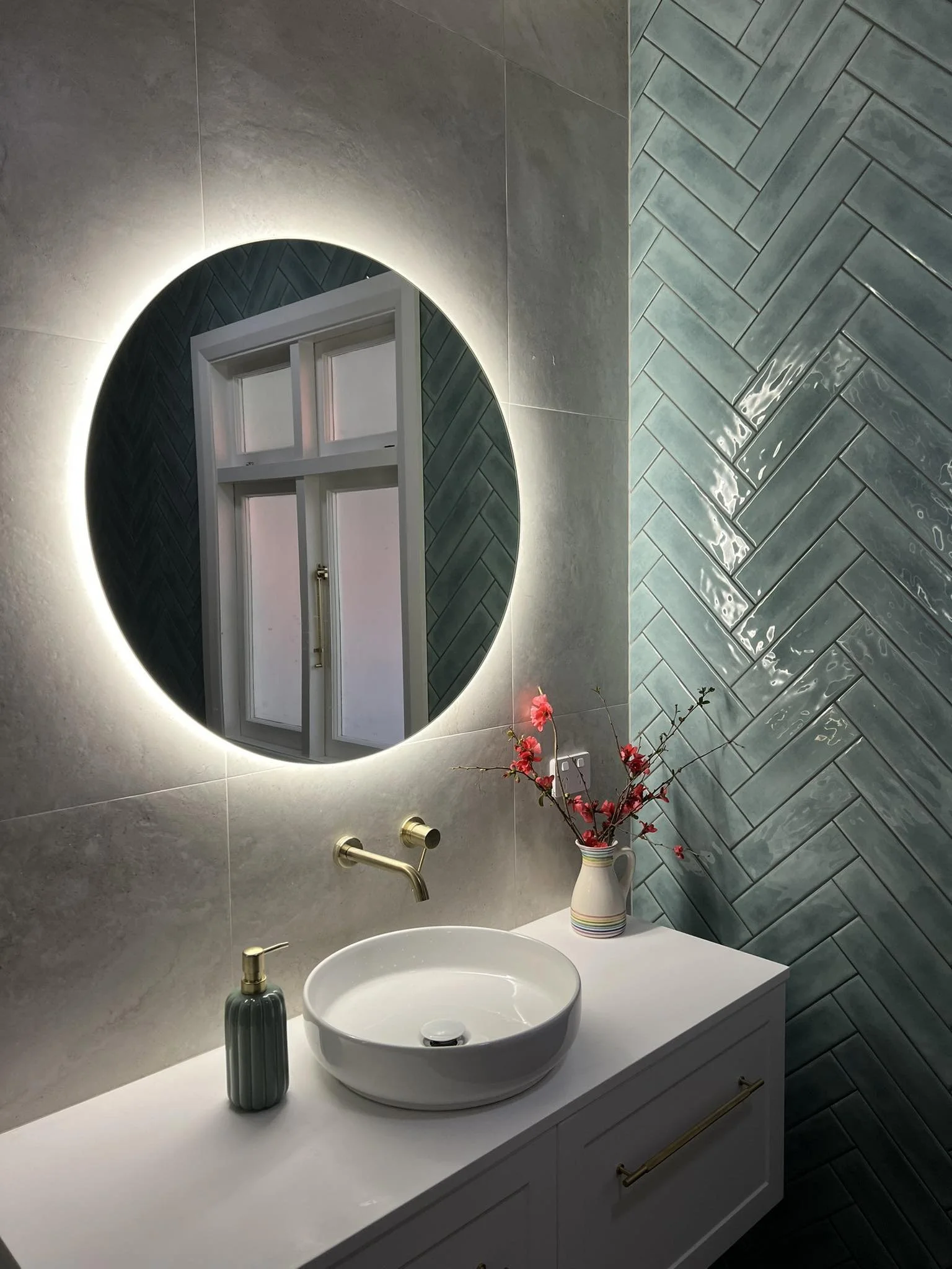 Modern bathroom with illuminated round mirror, white vanity with a vessel sink, gold faucet, soap dispenser, decorative vase with pink flowers, gray tiles, and a window reflected in the mirror.