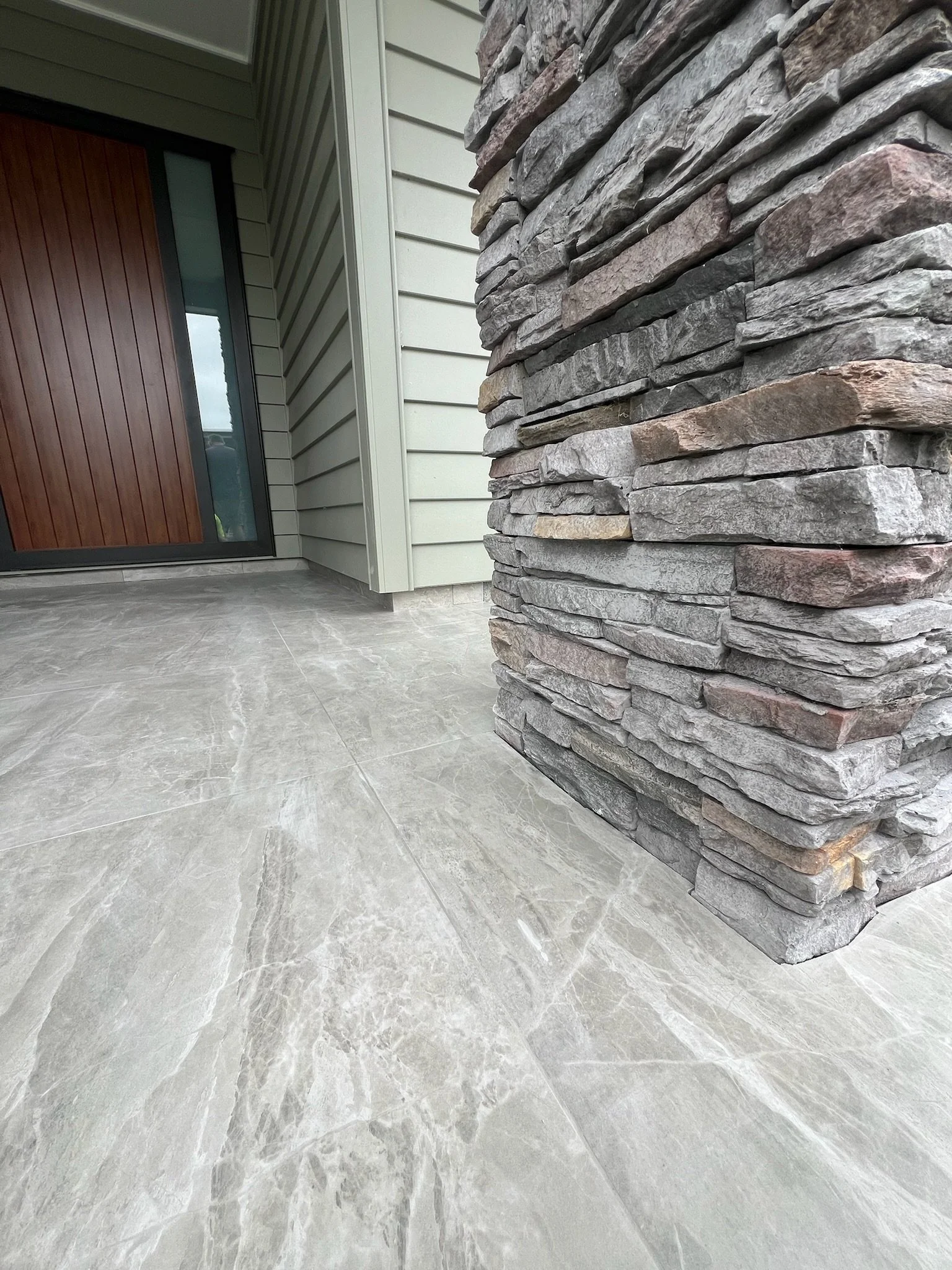 Close-up of a stone pillar next to a house entrance with grey tiled flooring and a green siding exterior.